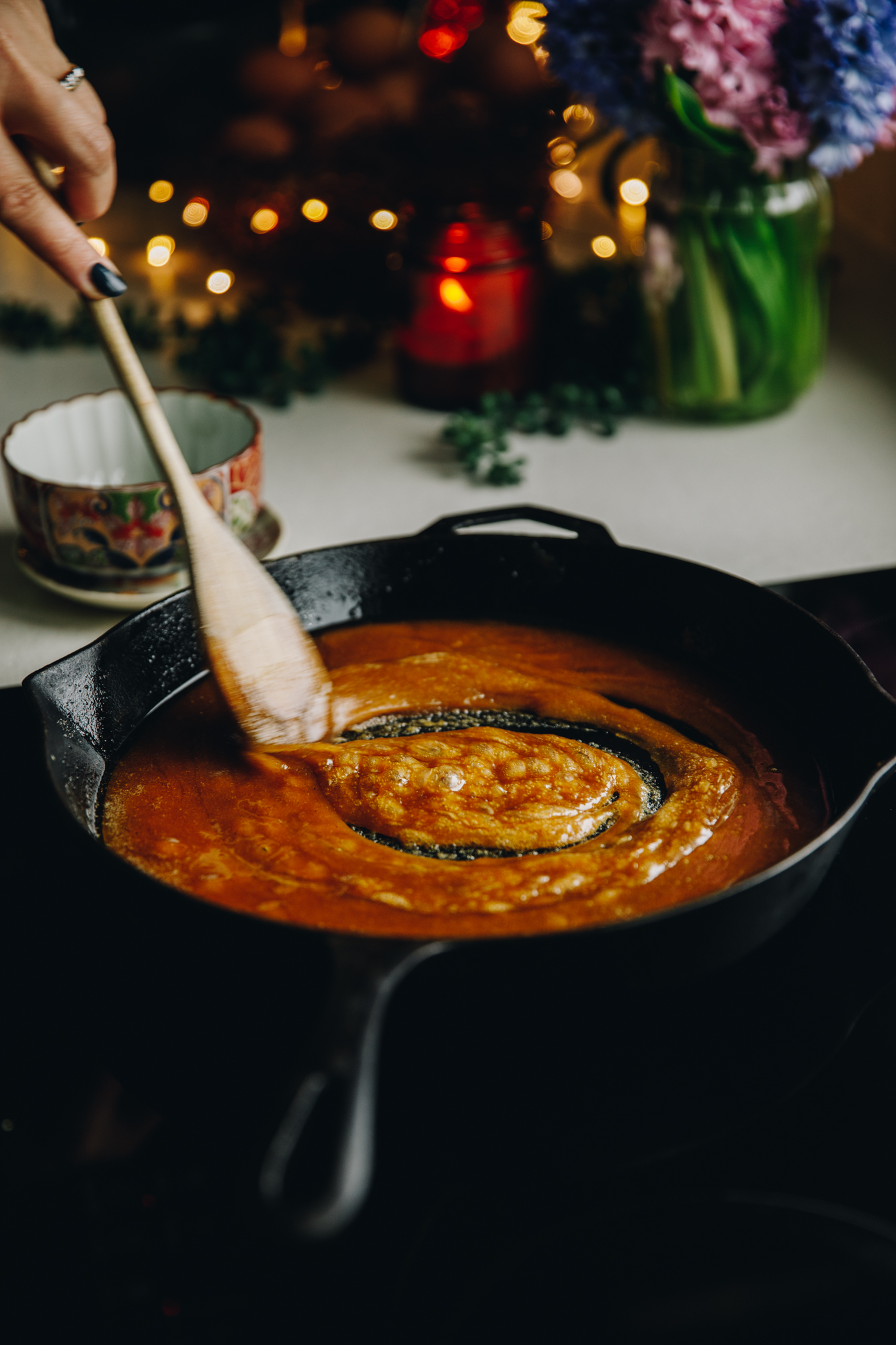 On a stove sits a cast iron pan with bubbly easy caramel sauce in it. A wooden spoon is stirring it as it bubbles. There is flowers and a candle on the bench behind it.