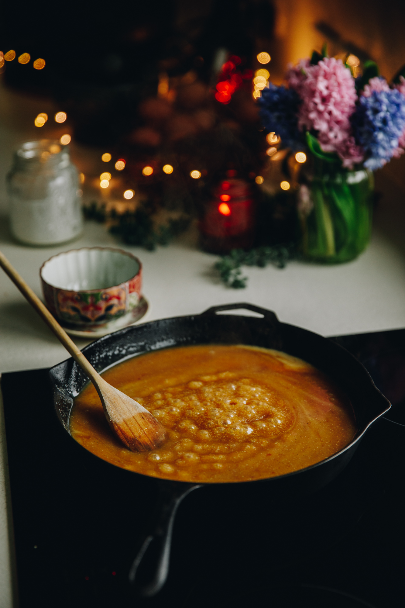 On a stove sits a cast iron pan with bubbly easy caramel sauce in it. A wooden spoon is stirring it as it bubbles. There is flowers and a candle on the bench behind it.