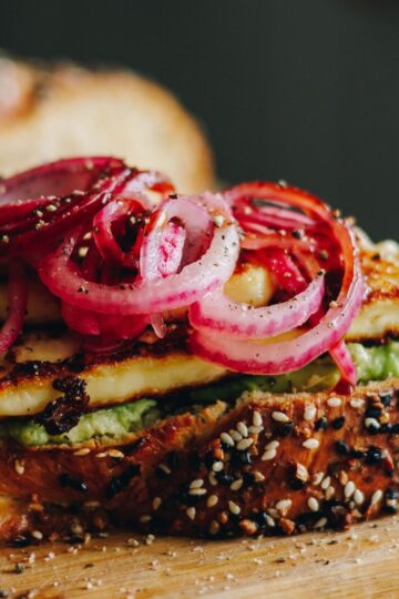 Close up shot of freshly baked bread with bagel season on top shows layers on avocado, grilled halloumi cheese and red pickled onions.