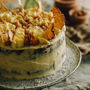 A whole sticky caramel date cake is on a cake stand. It is covered in buttercream and is decorated with buttercream squiggles and dried apples crisps.