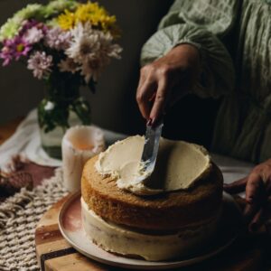 A two layered vanilla cake is being decorated in with simple cream cheese frosting that is being spread on to the top of the cake. It is on a wooden board with flowers in the background.