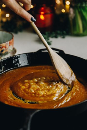 In a black cast iron pan is easy caramel sauce simmering, it is being stirred with a wooden spoon.