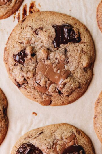 Freshly baked brown butter chocolate cookies are on brown baking paper. The flay lay shot shows a close up of a cookie with melty dark and milk chocolate chunks on it.
