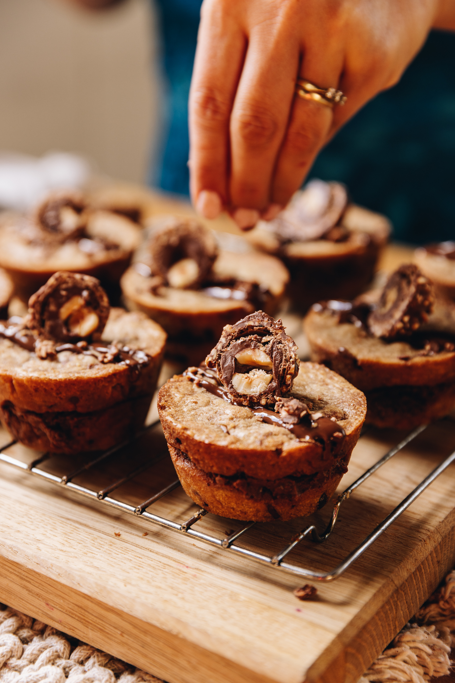 Baked gooey cookie pies are on a wire rack, on a wooden board. They are drizzled with chocolate, a forerro rocher is on top of each one and a hand is sprinkling chopped nuts on top.