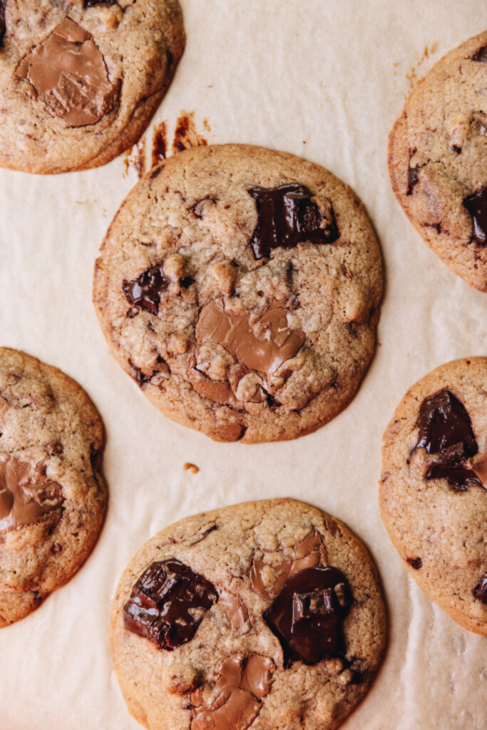 Freshly baked brown butter chocolate cookies are on brown baking paper. The flay lay shot shows a close up of a cookie with melty dark and milk chocolate chunks on it.