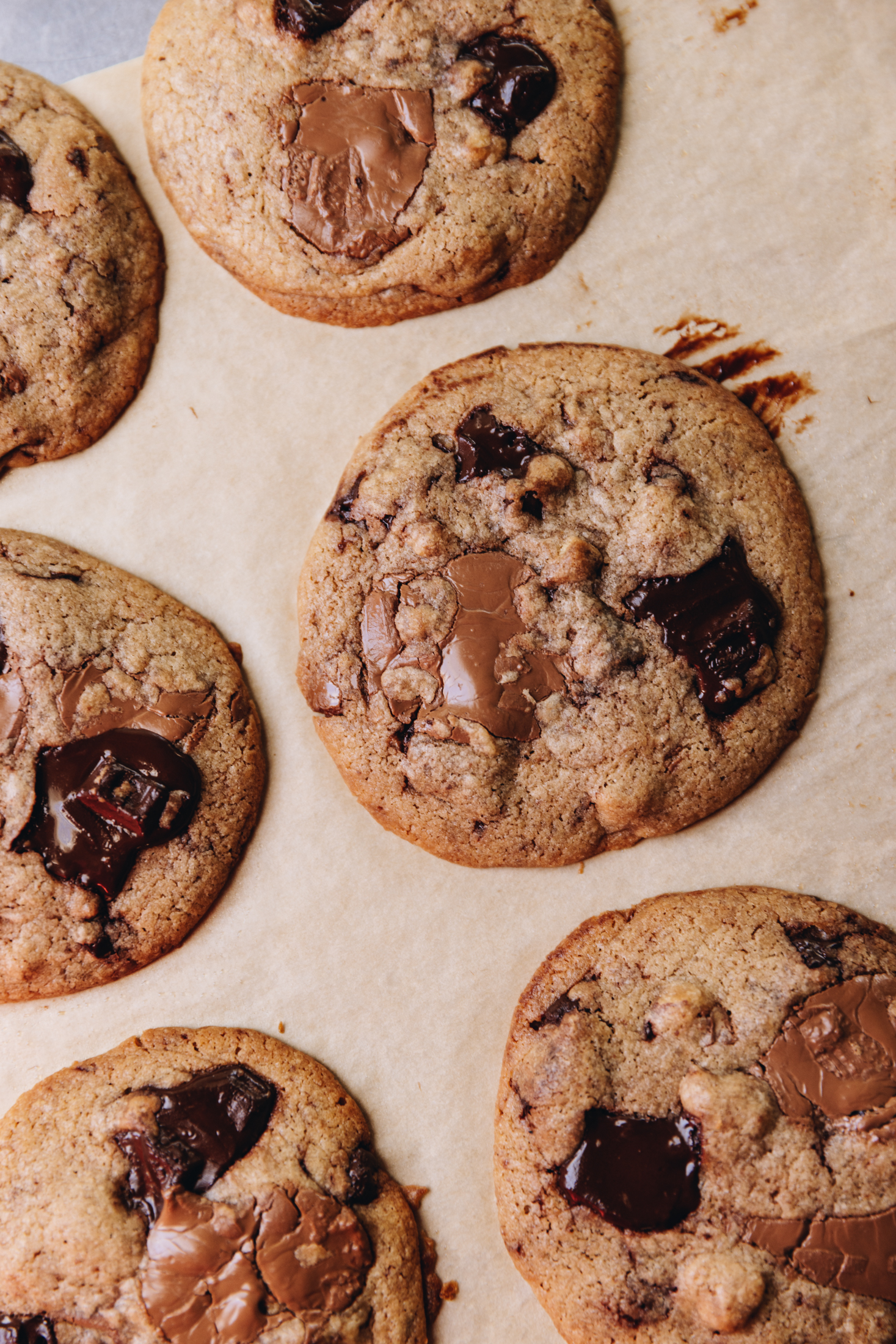 Freshly baked brown butter chocolate cookies are on brown baking paper. The flay lay shot shows a close up of a cookie with melty dark and milk chocolate chunks on it.
