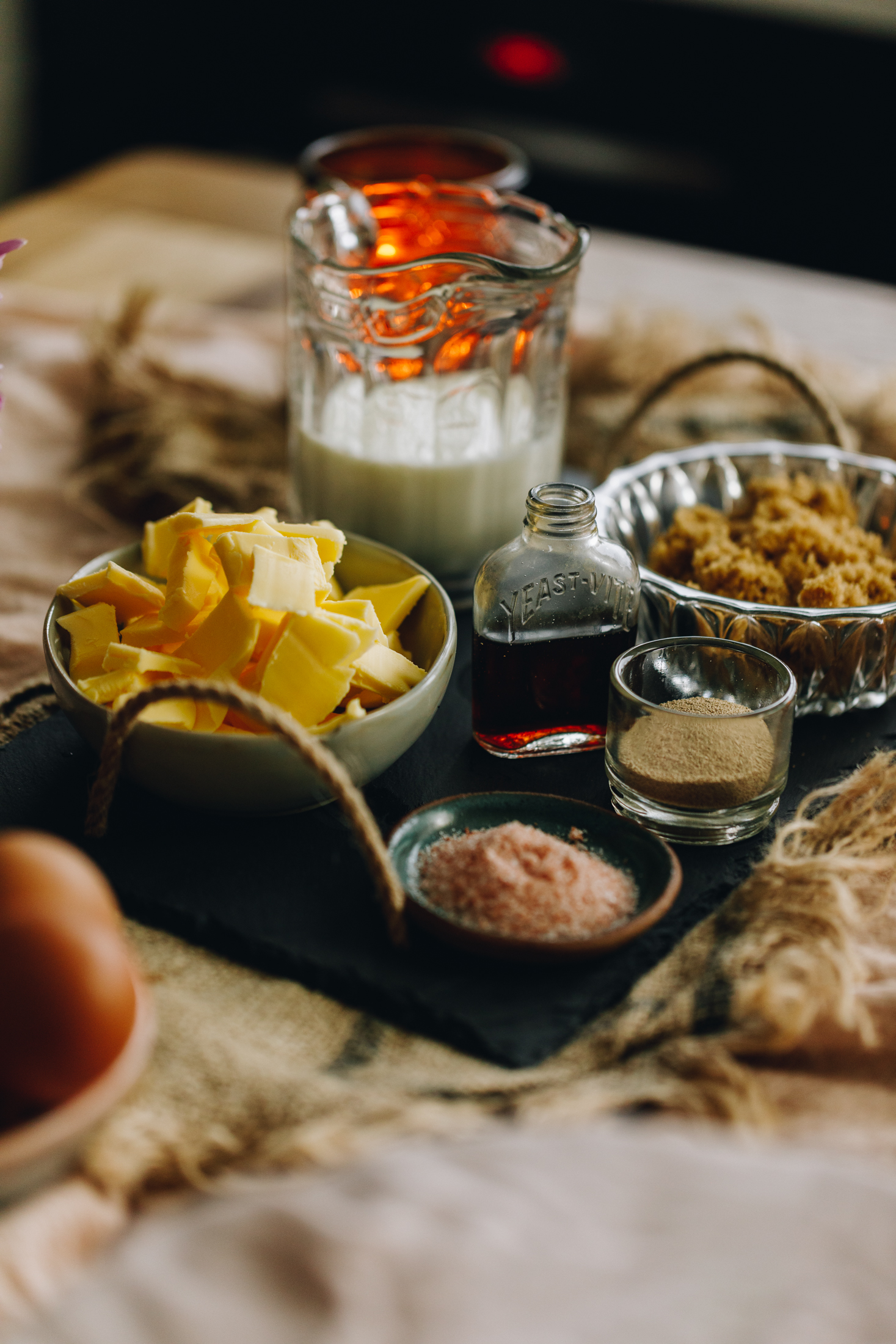 A small black tray with rope handles ia on a table lined with natural fabrics. On the tray is vintage bowls, jars and jugs with salt, yeast, brown sugar, butter and milk in it. In the background an orange candle is burning. Eggs are in the forefront of the photo.