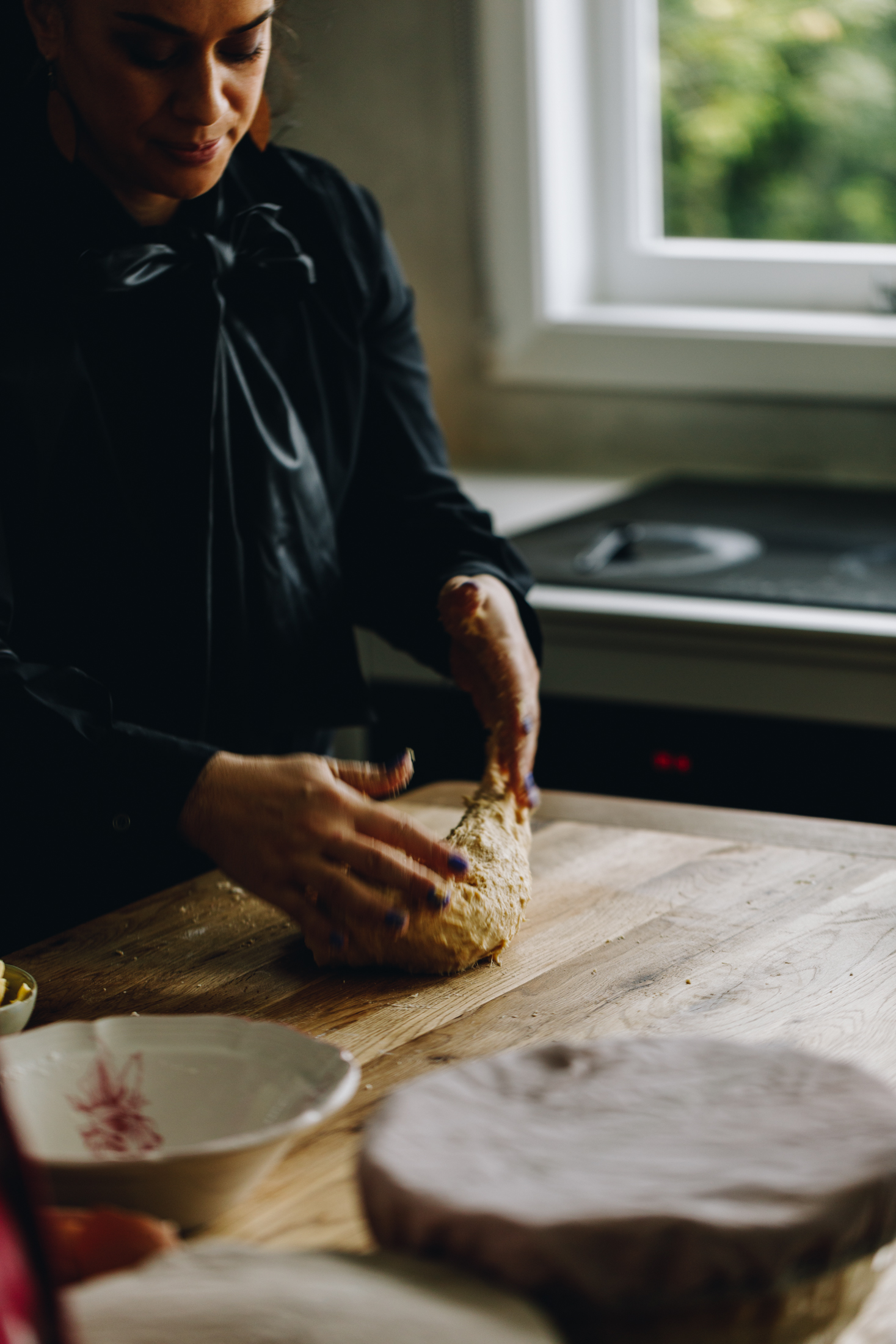 Naomi Toilalo is standing behind a wooden table kneading brioche dough. A window with a green tree is in the background and a bench with a black stovetop. Bowl are seen in the foreground. 