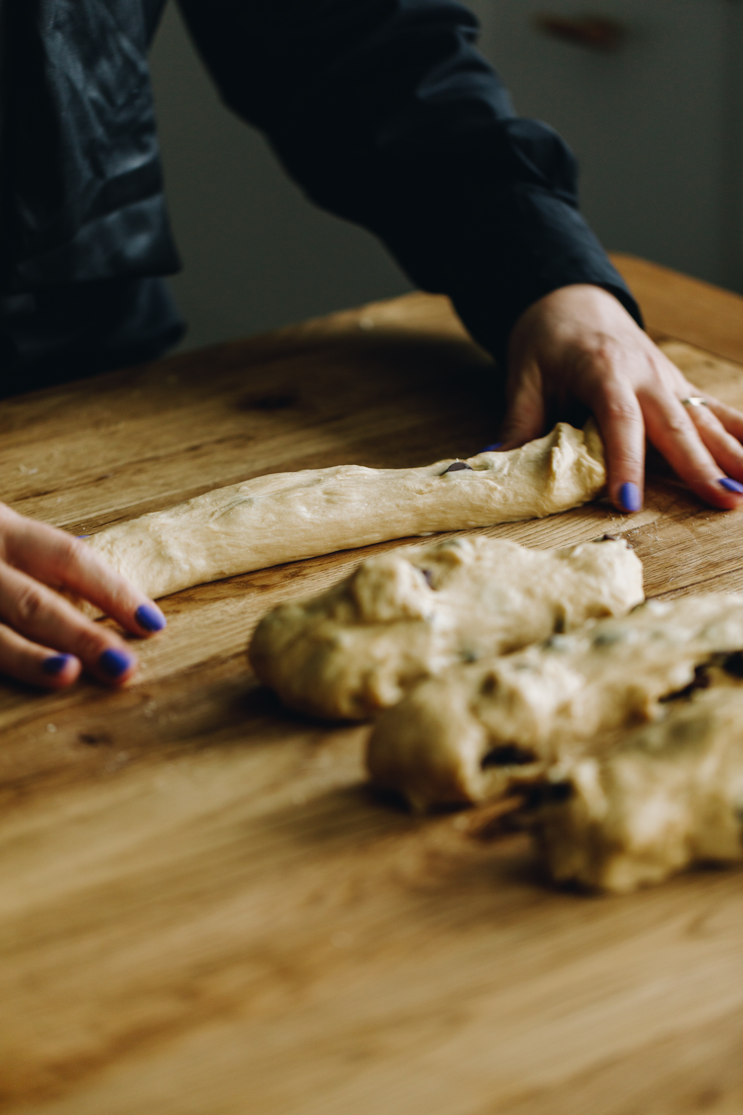 On a wooden table are four pieces of dough and one of them is being rolled in to a rope with Naomi's hands. 