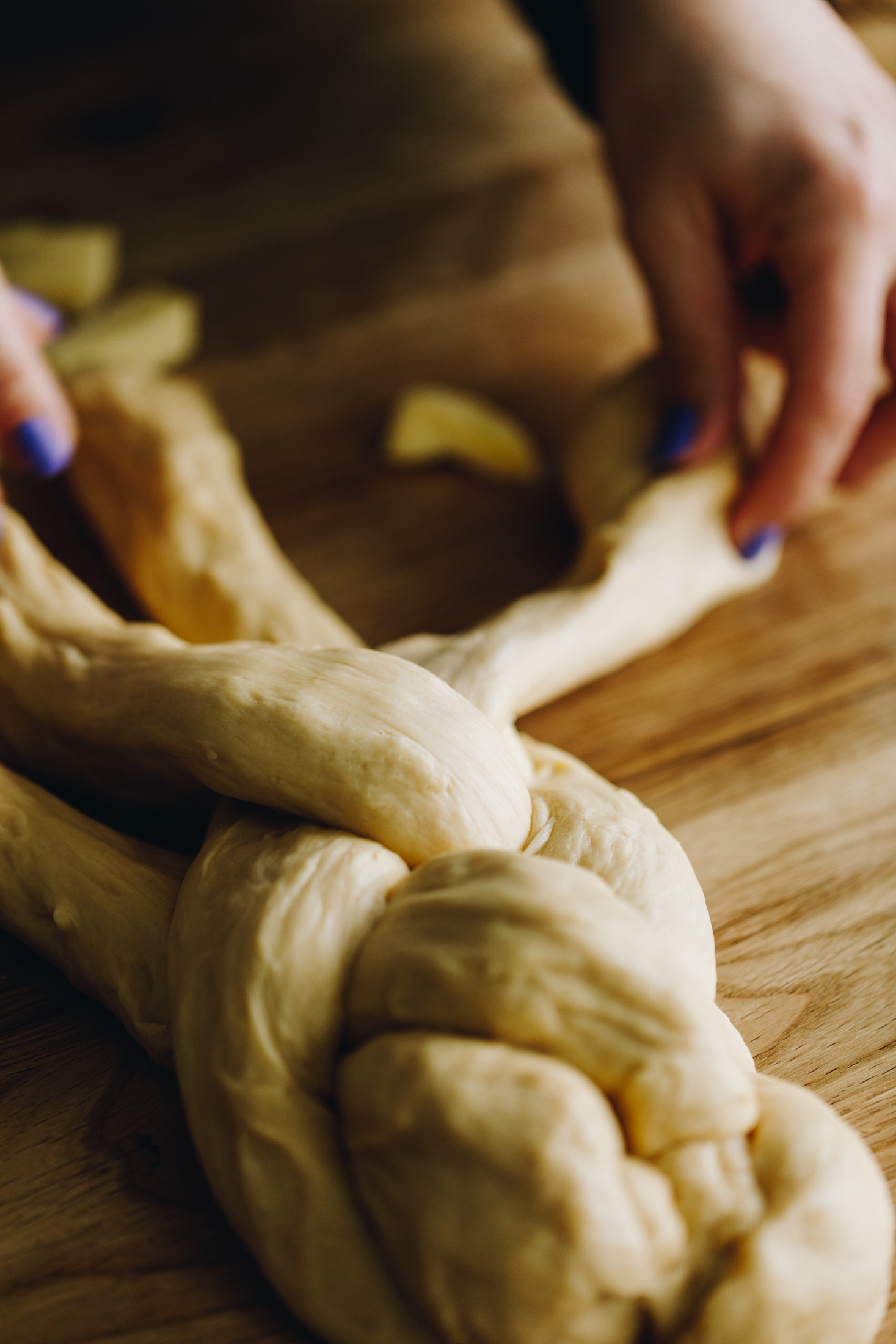 On a wooden table four strands of brioche bread dough are being braided together with Naomi's hands. 