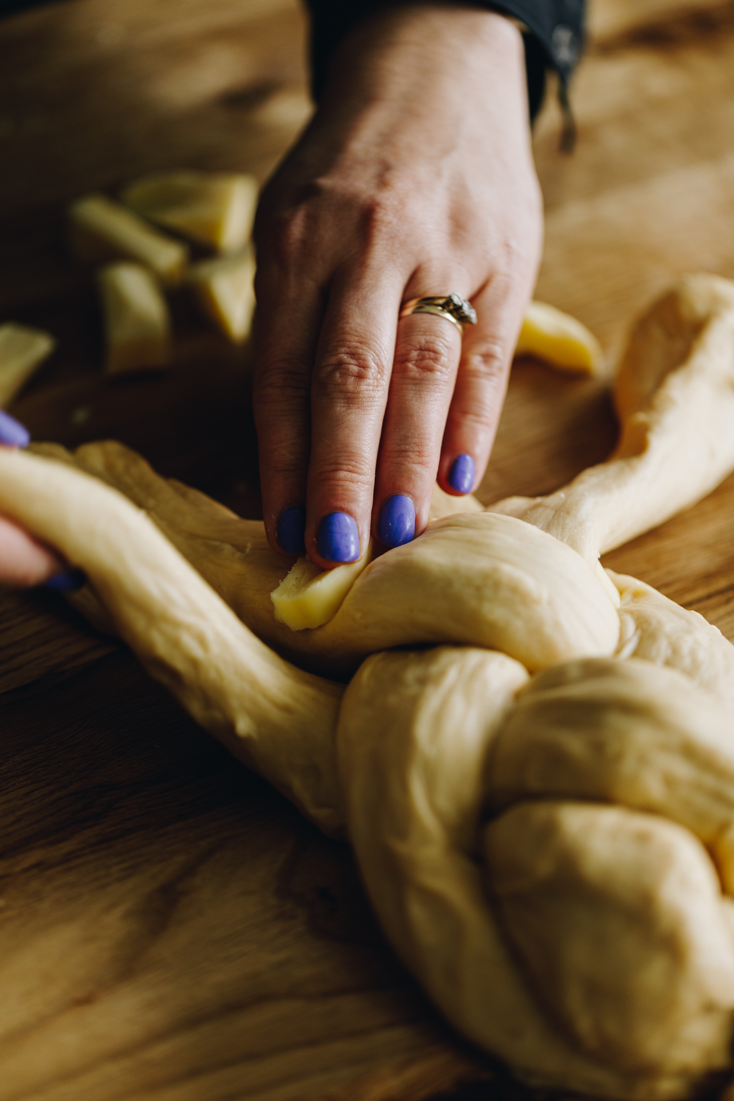 On a wooden table four strands of brioche bread dough are being braided together with Naomi's hands. A piece of cheese is being placed one to one of the strands and another strand is beginning to move over it. 