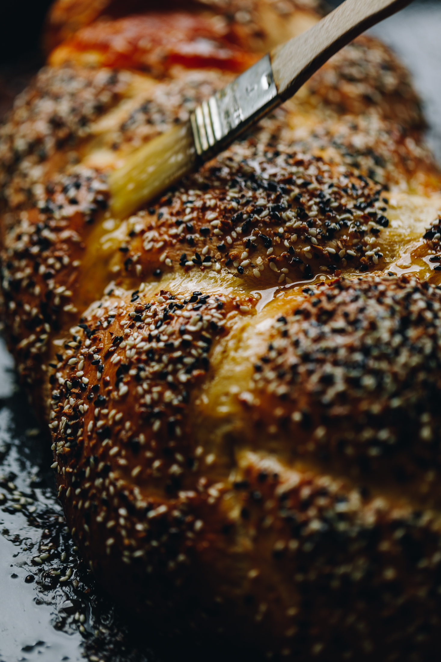 A Savoury braided brioche bread loaf sits on a black oven tray. It is golden and bagel season is sprinkled over it. It is being brushed with melted butter making the loaf super golden.