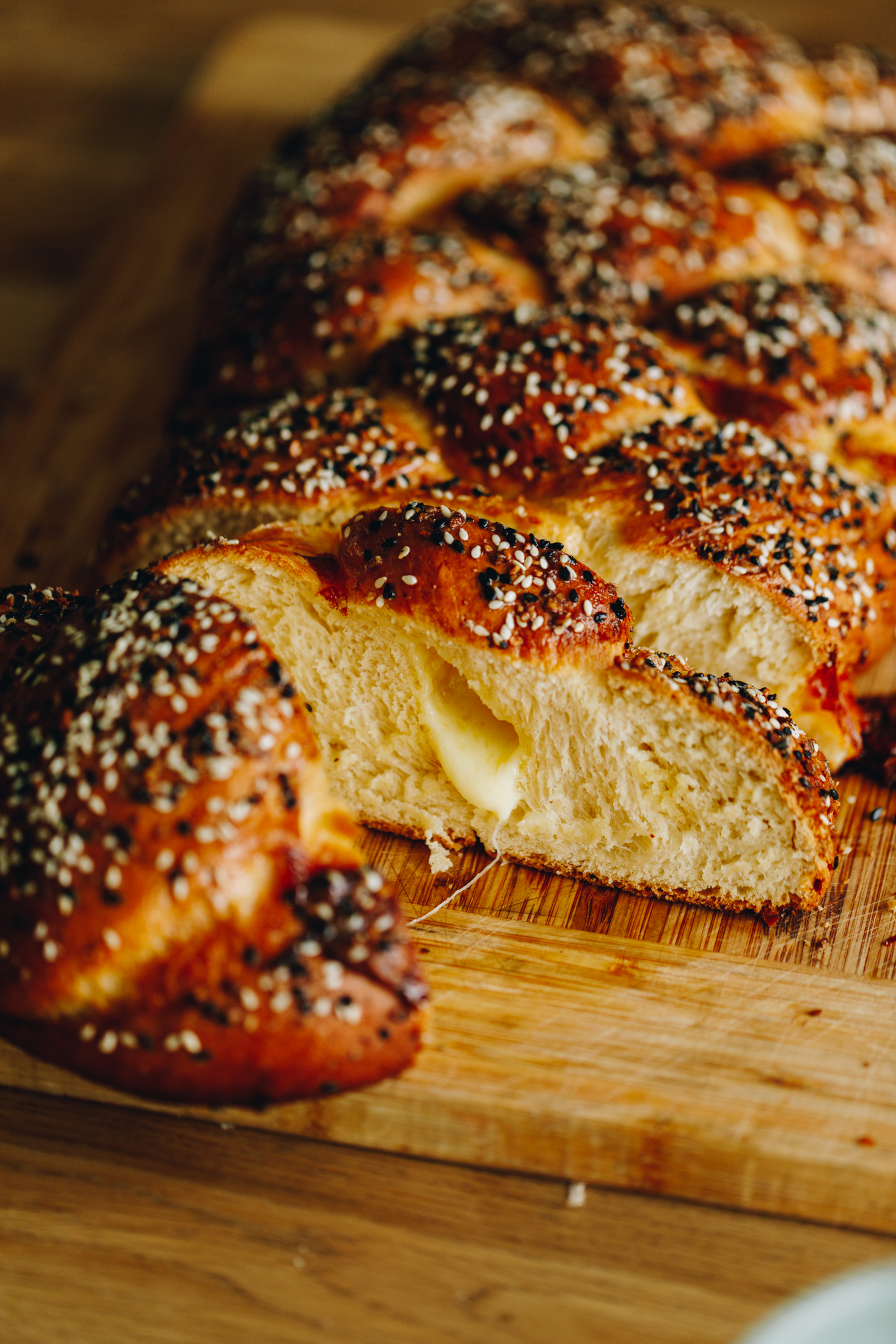 On a wooden table is a wooden chopping board. On the board sits a savoury braided brioche bread that has been cut open to reveal cheese oozing out of the centre. It is golden on top and has been sprinkled with bagel mix on top of the bread.