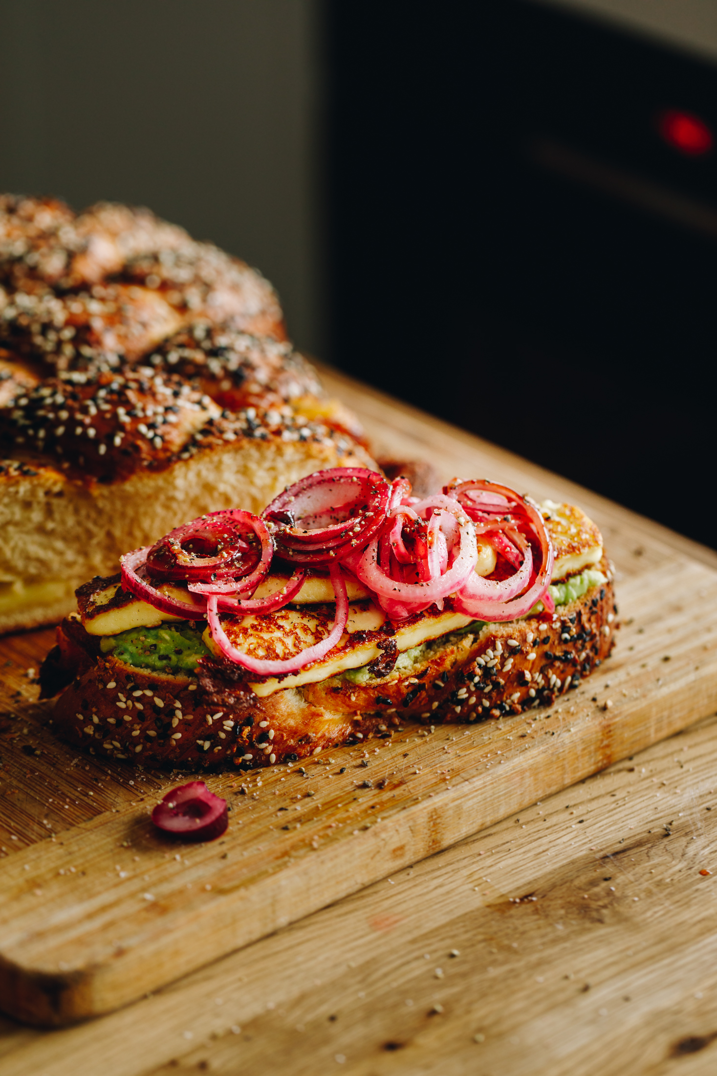 A wooden board sits on a wooden table. On the board is a freshly baked Savoury braided brioche bread loaf that has been cut open. In front of it is a slice of the bread with avocado smeared on to it with grilled halloumi pieces and piled with red pickled onions.  