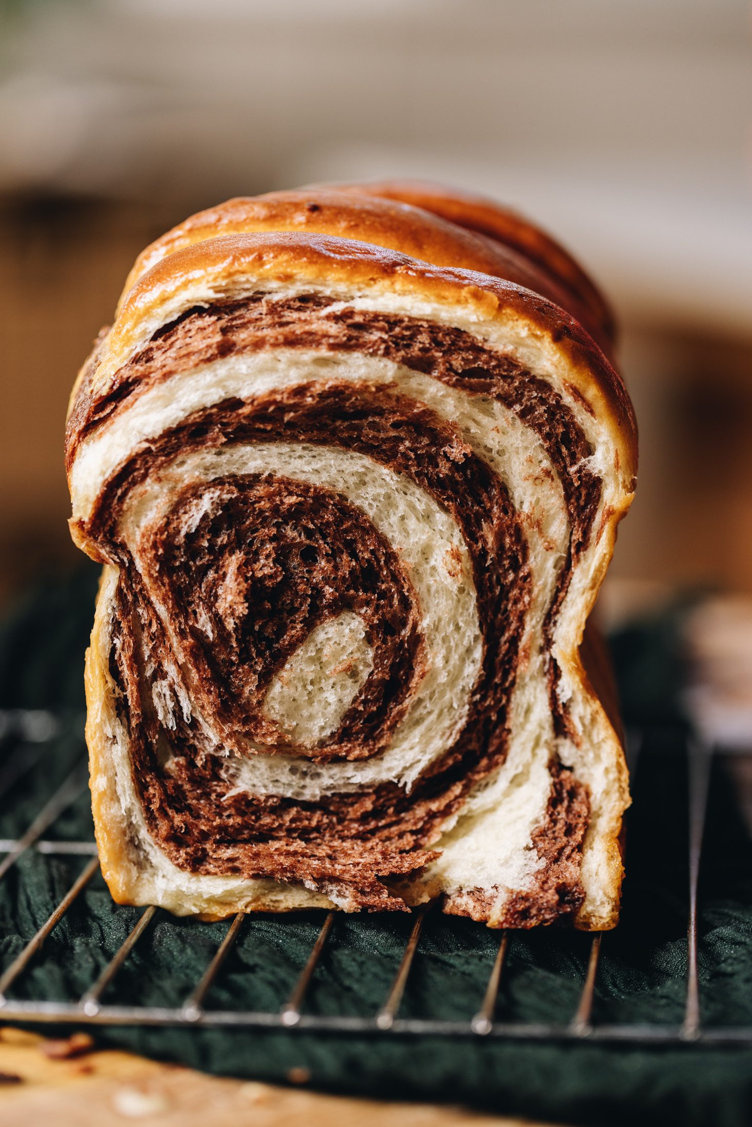 A freshly baked loaf of chocolate swirl bread is on a cooling rack which sits on a wooden table, with a green cloth underneath it. Is has been sliced open to reveal the chocolate and vanilla swirl pattern inside the bread loaf. 