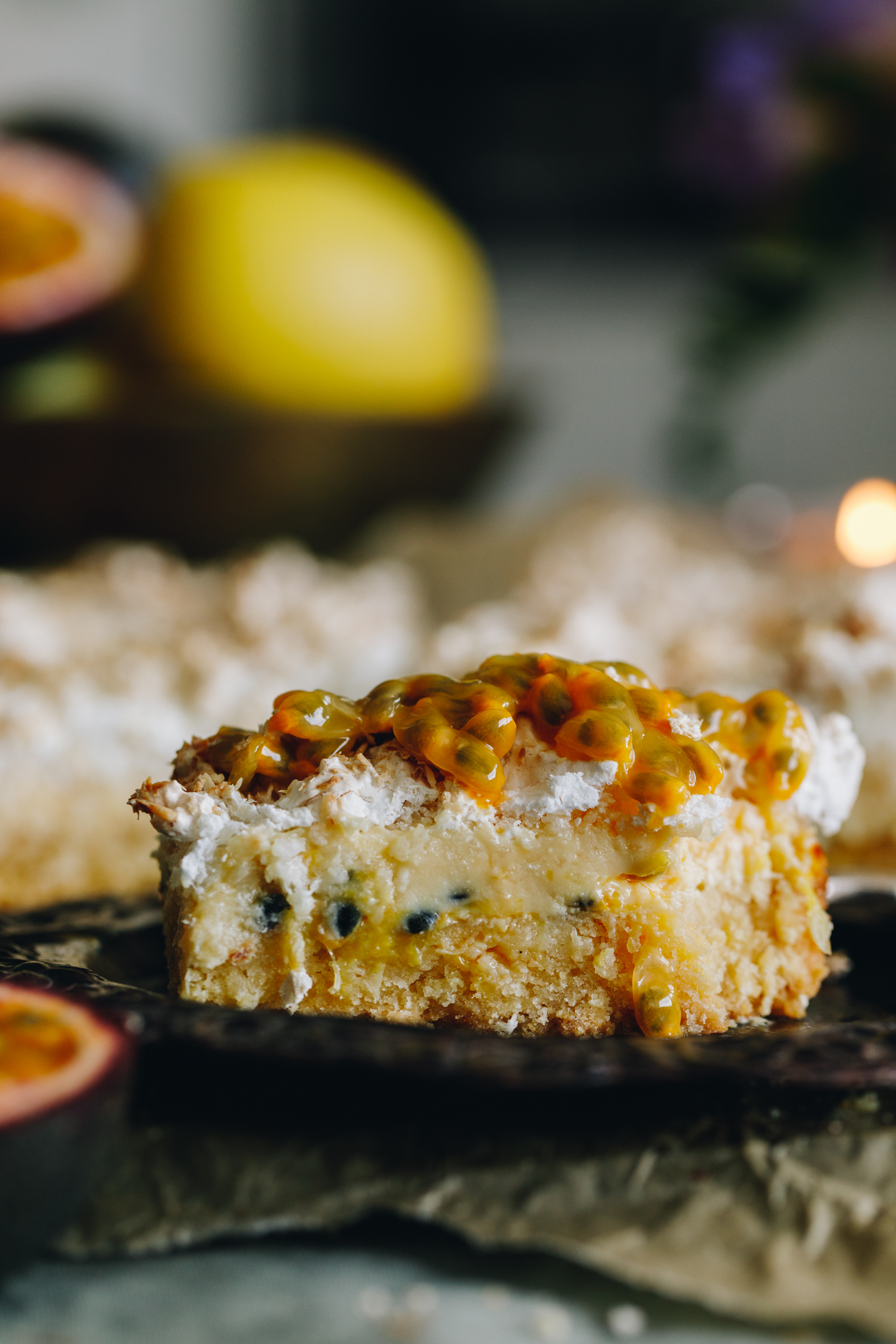 A slice of passionfruit lemon meringue slice is on a small black plate that is on layers on green cloths. The close shot shows the buttery base, creamy passionfruit centre and the coconut meringue on top. It has fresh passionfruit on top. Lemons are in a bowl blurred in the background. 