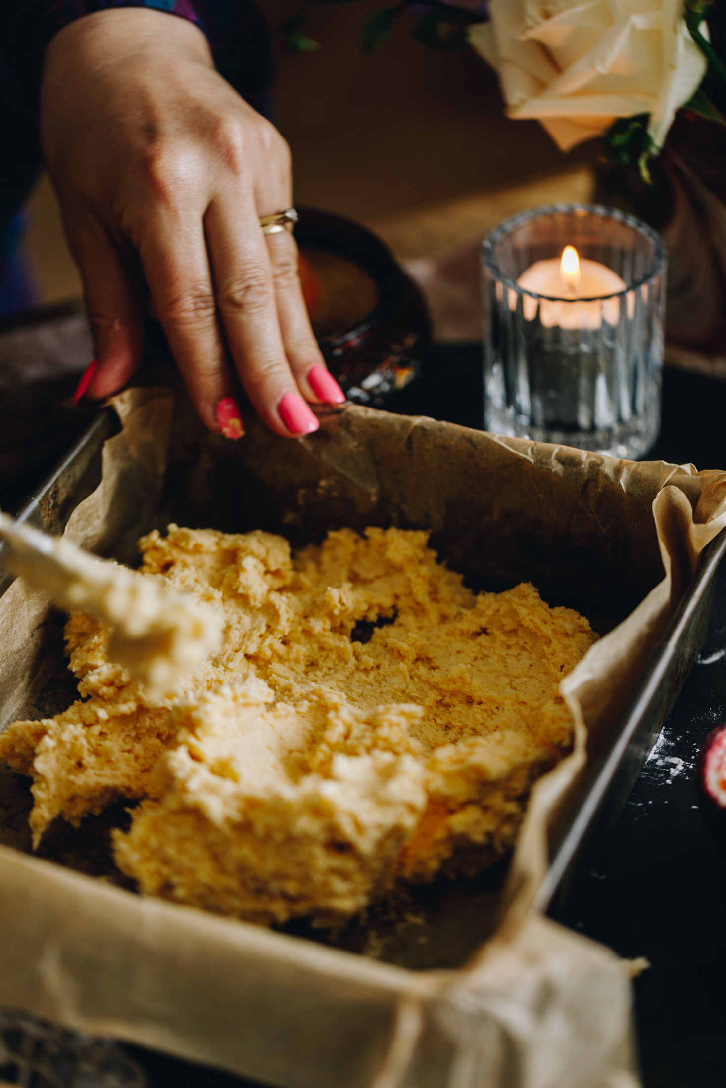 A vintage baking tin is lined with brown baking paper and is sitting on a black stone. In the tray is a coconut and whipped cream butter mixture which is being spread in to the tin. A small glass with a silver candle burns behind it and a white rose is behind it. 