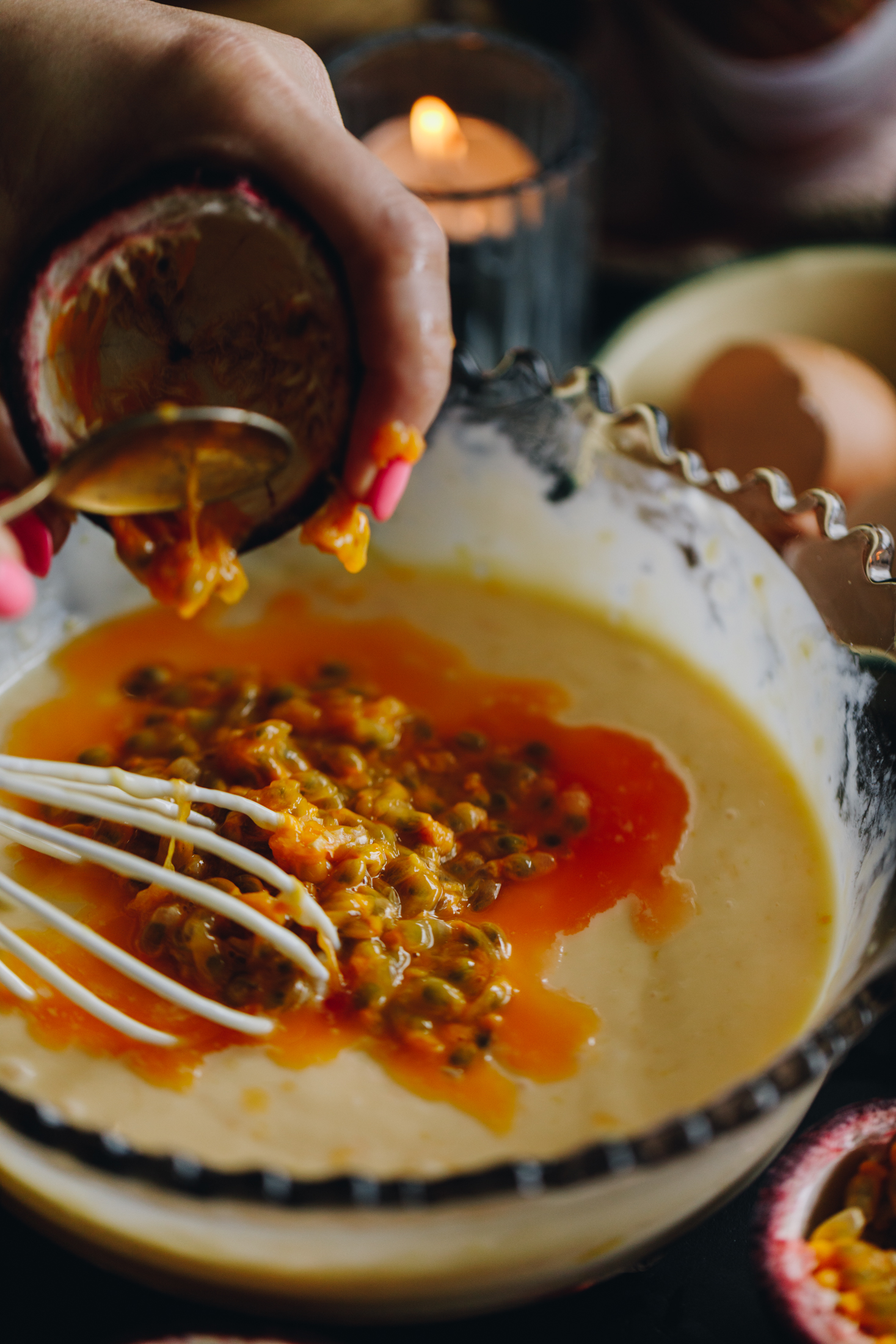 A purple vintage bowl has a creamy mixture in it and Naomi is emptying fresh passionfruit pulp in to the bowl with a spoon. A white wire whisk is sitting in the mixture. Broken egg shells are in a bowl in the background along with a candle burning in a small glass.