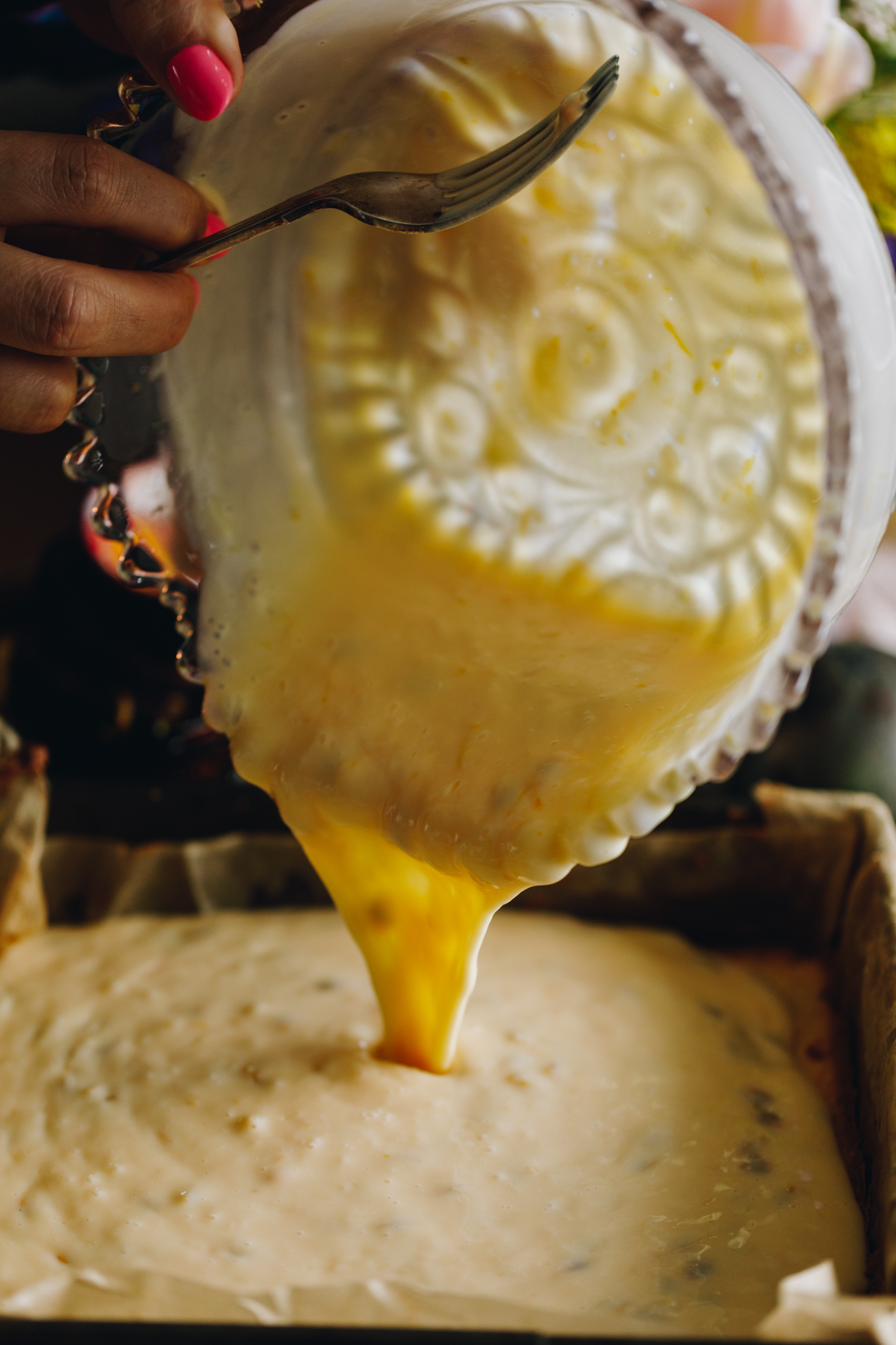 A vintage baking tin is lined with brown baking paper. A purple vintage bowl is pouring the passionfruit custard mixture in the tin. The mixture is still pouring in. Naom is holding a fork in the bowl too. 