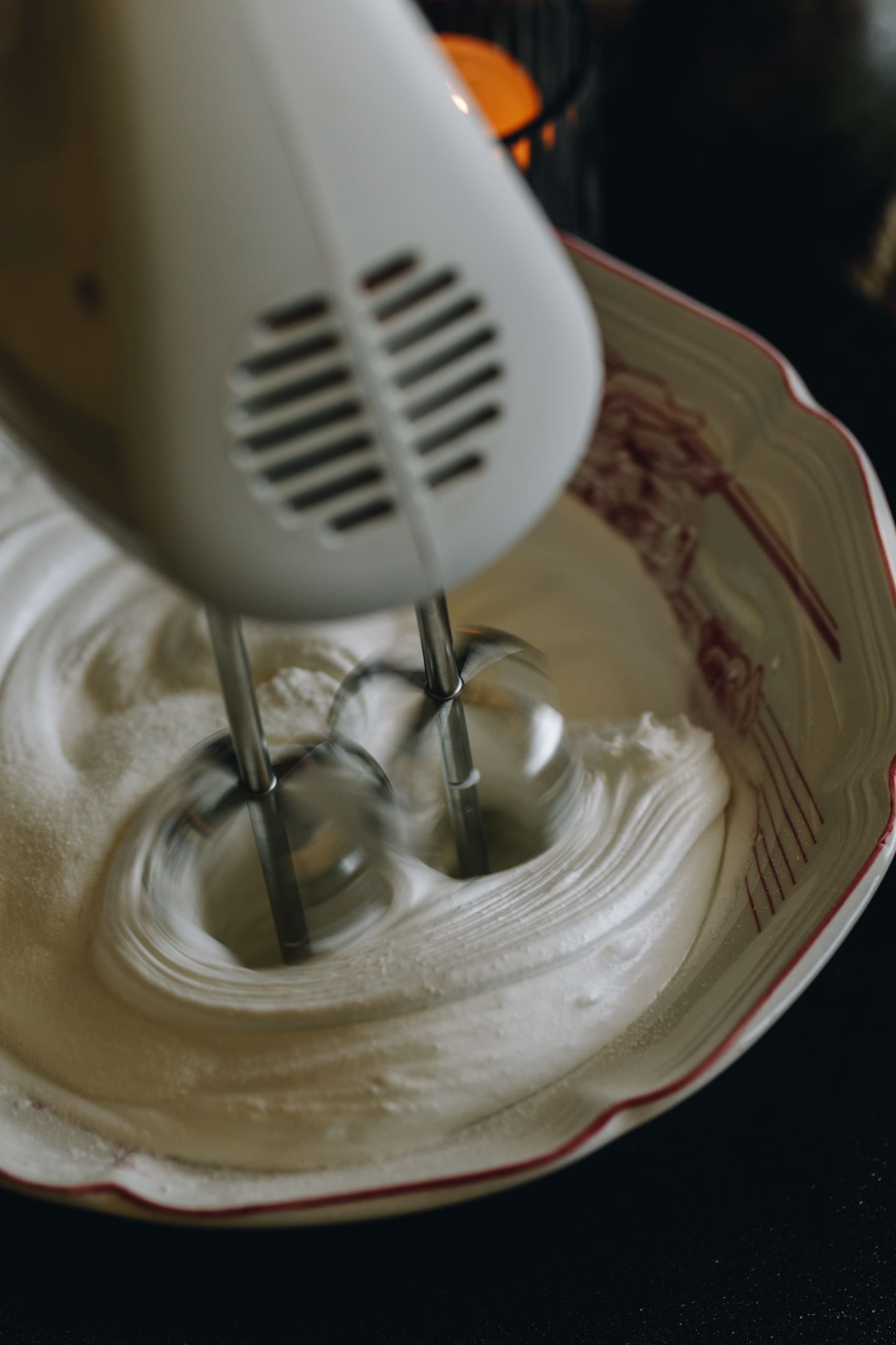 A vintage white bowl with pink flowers on it has meringue inside it. It is being whipped with a white hand mixer. 