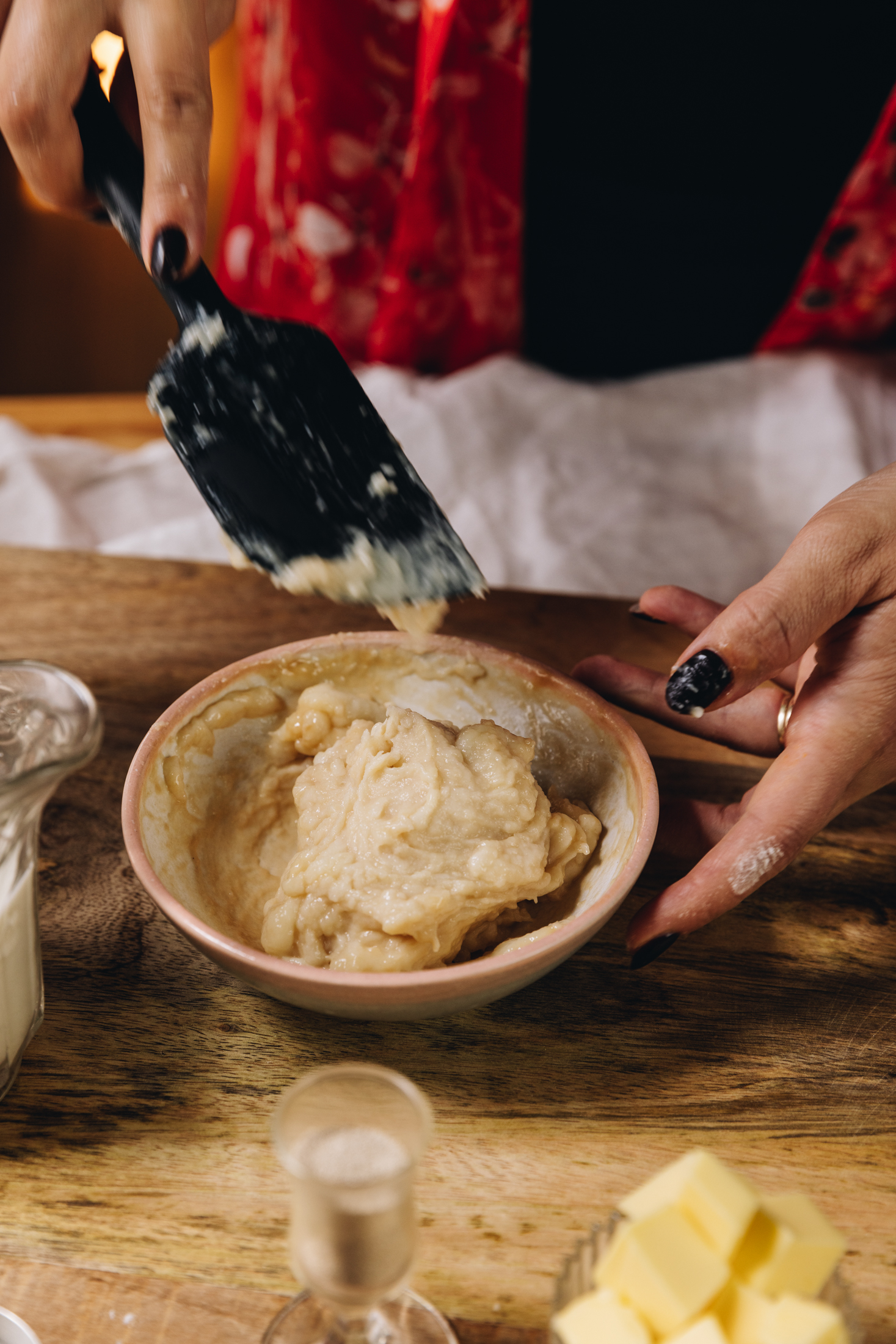 A small pink bowl sits on a wooden table with tangzhong in it. Naomi has a black spatula hovering above it. A glass jug is to the side of Fram, a small glass and cubed butter is in front.