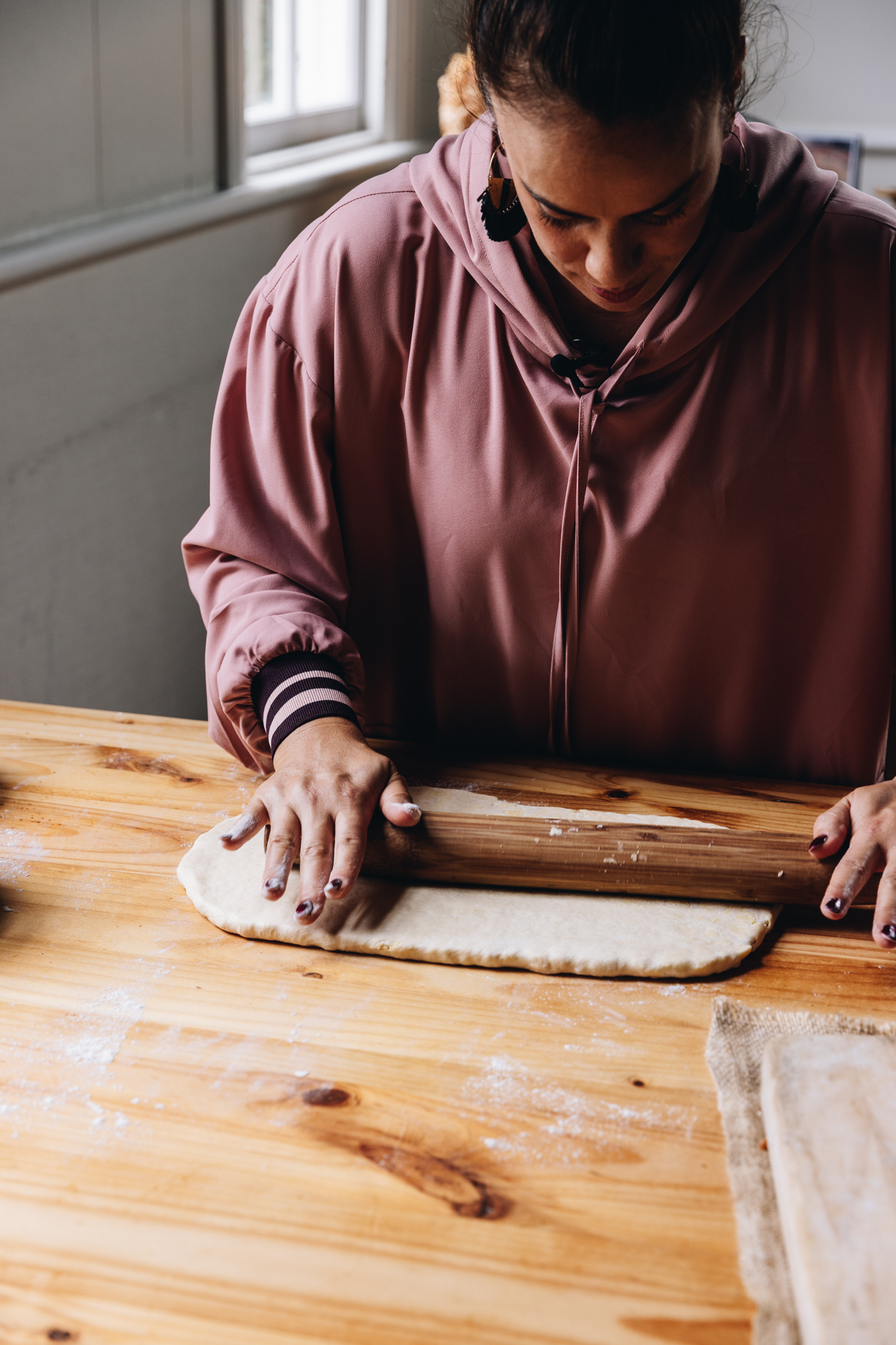 Naomi Toilalo is rolling out a dough on to a lightly floured wooden table with a wooden rolling pin. 