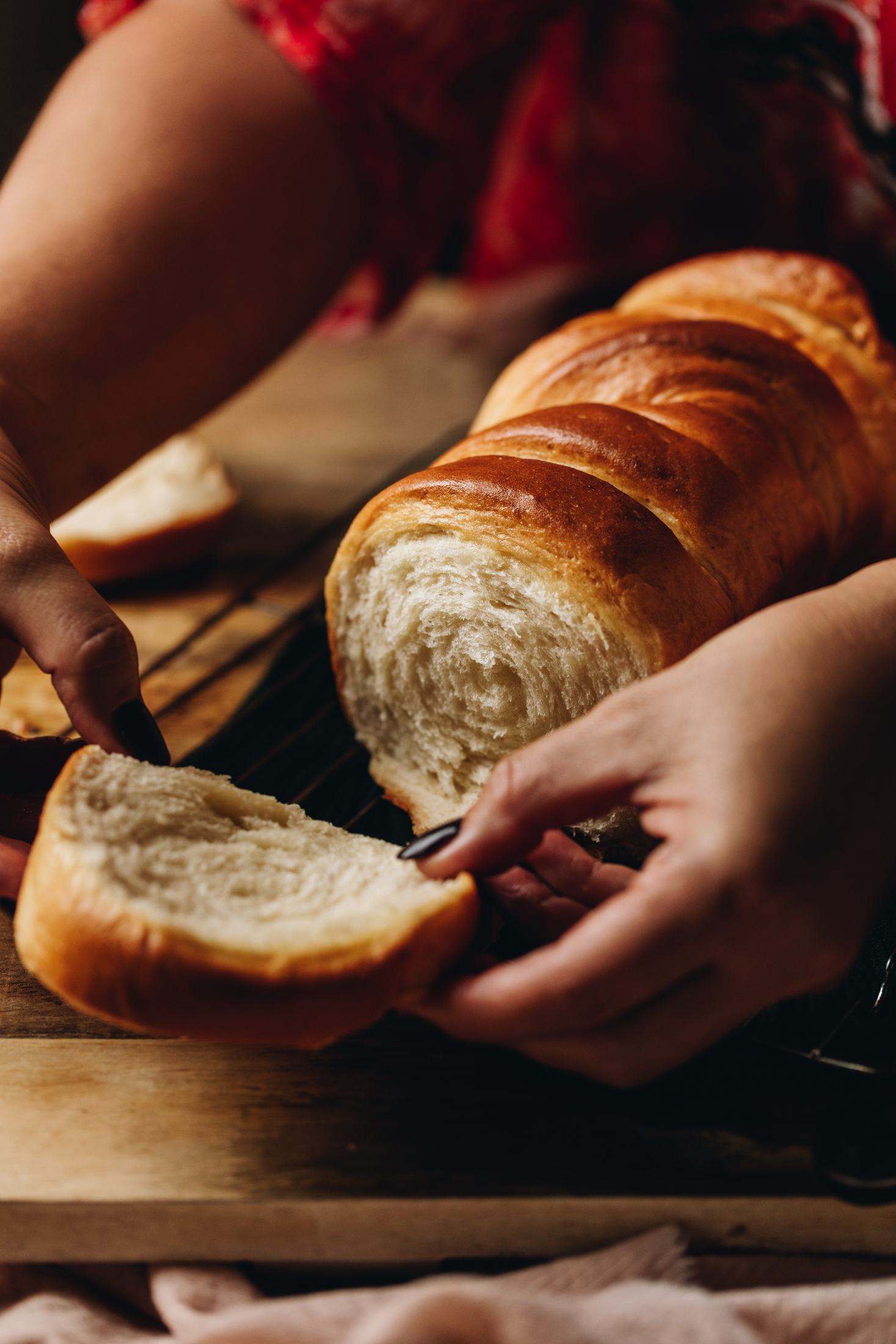 A freshly baked milk bread loaf sits on a wooden board on a pink tablecloth. Naomi is pulling a piece off the loaf which reveals the spiral bread and soft texture. 