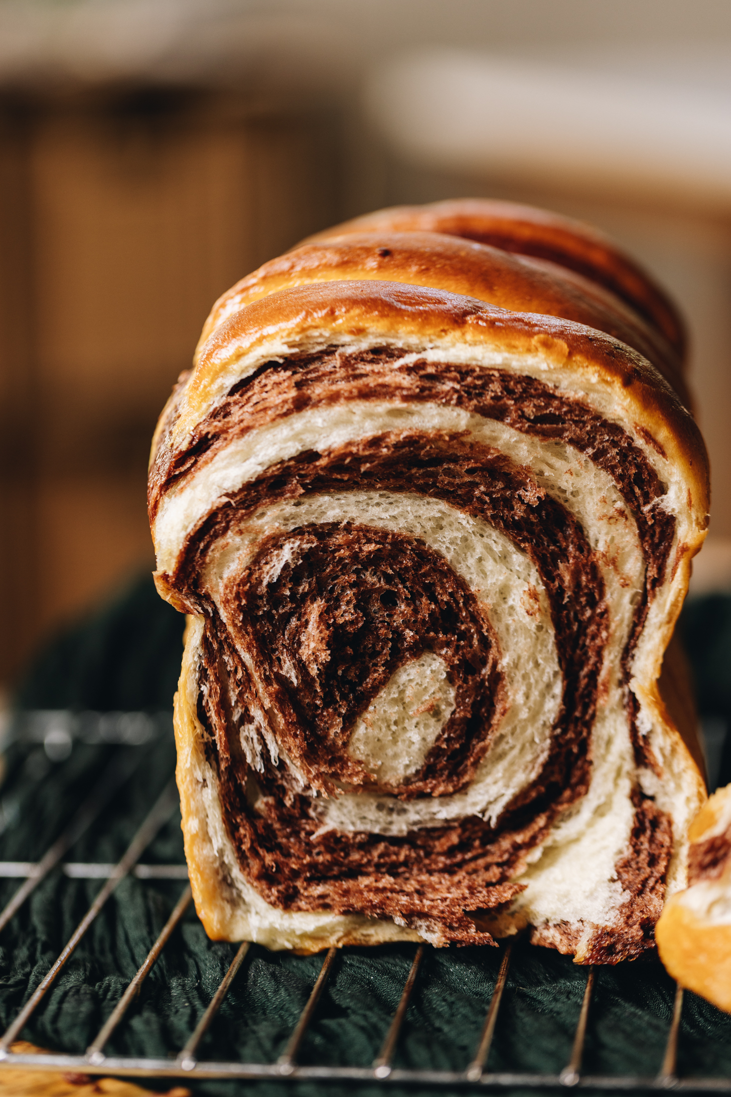 A freshly baked chocolate swirl loaf has been cut open revealing the swirl pattern in the loaf. It is on a wire cooling rack on a green cloth, on top of a wooden table. 