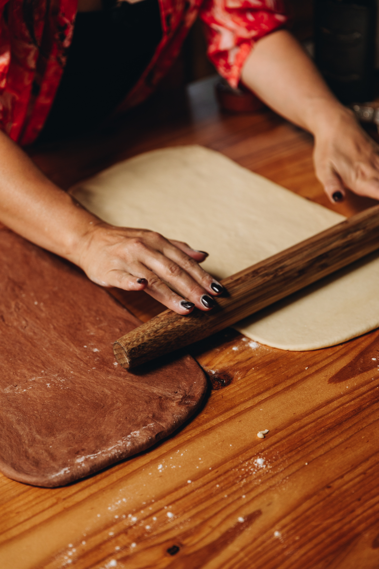 A chocolate dough is rolled out and a plain dough is being rolled out on a wooden table.