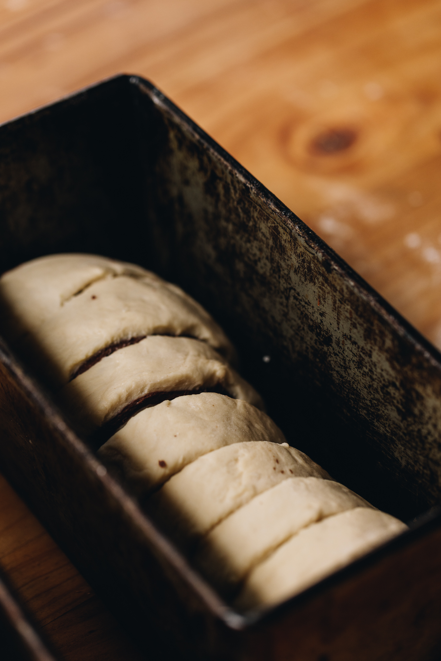 Unbaked slices of the chocolate swirl bread loaf are in a rustic loaf tin that sits on a wooden table.