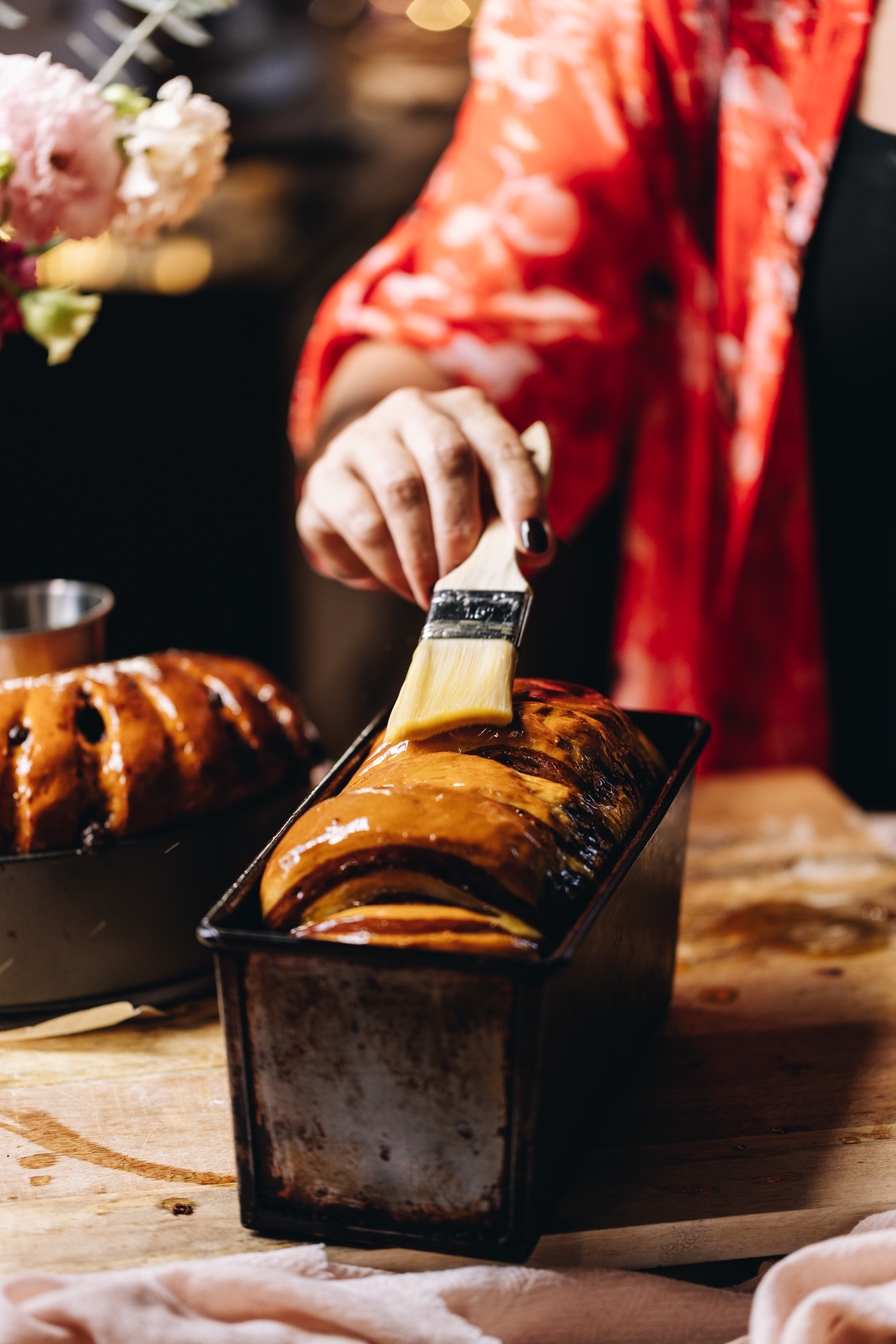A freshly baked chocolate swirl bread loaf is being glazed with butter on a wooden table. Behind it is another baked loaf and peach flowers in a vase.