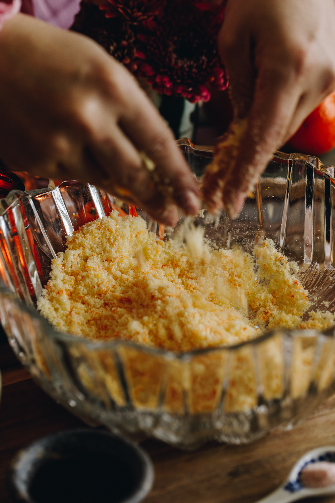On a wooden table sits a glass vintage bowl that has sugar and orange zest in it. Naomi is using her hands to rub the zest in to the sugar.
