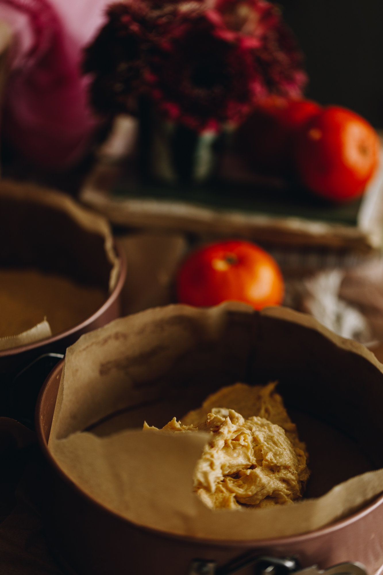 Two cake tins lined with baking paper are sitting on a wooden table. One has cake mixture in it. Oranges and flowers are in the background.