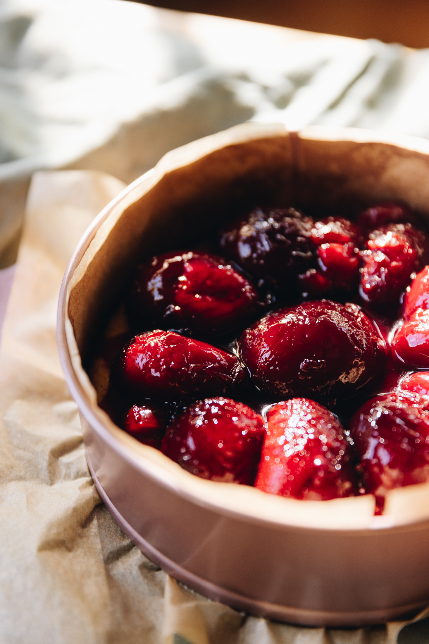 On cake tin sits a rose pink cake tin that is lined with brown baking paper. On the cake is halved black Doris plums. Under the tin is a soft green tablecloth.