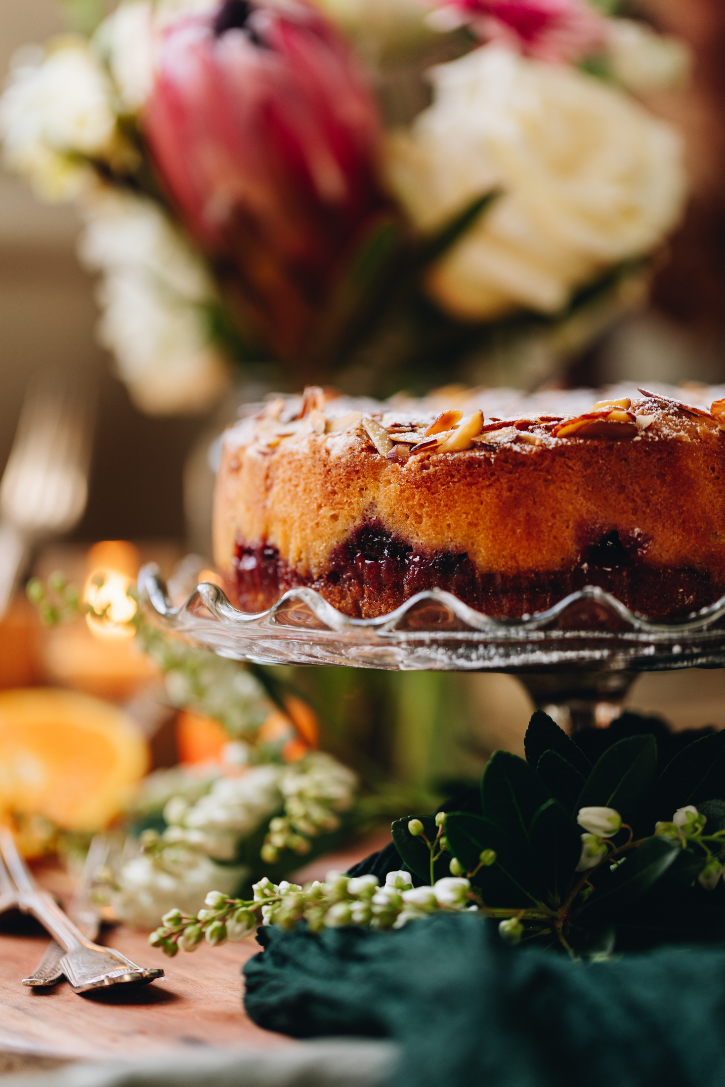 On a cake stand sits a freshly baked plum and almond cake. Behind it is a large bunch of flowers with cut oranges and greenery around it, all sitting on a wooden table.