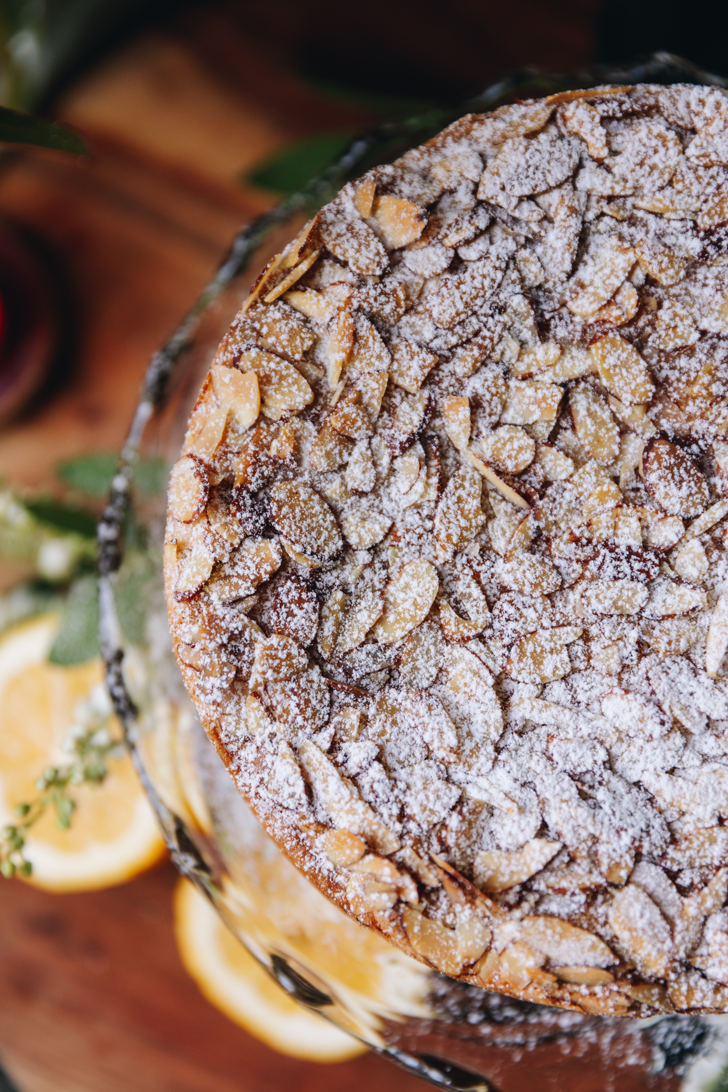 A top shot of a whole plum and almond cake shows the sliced almond topping with a dusting of icing sugar. Sliced lemons and greenery are seen on the wooden table underneath.