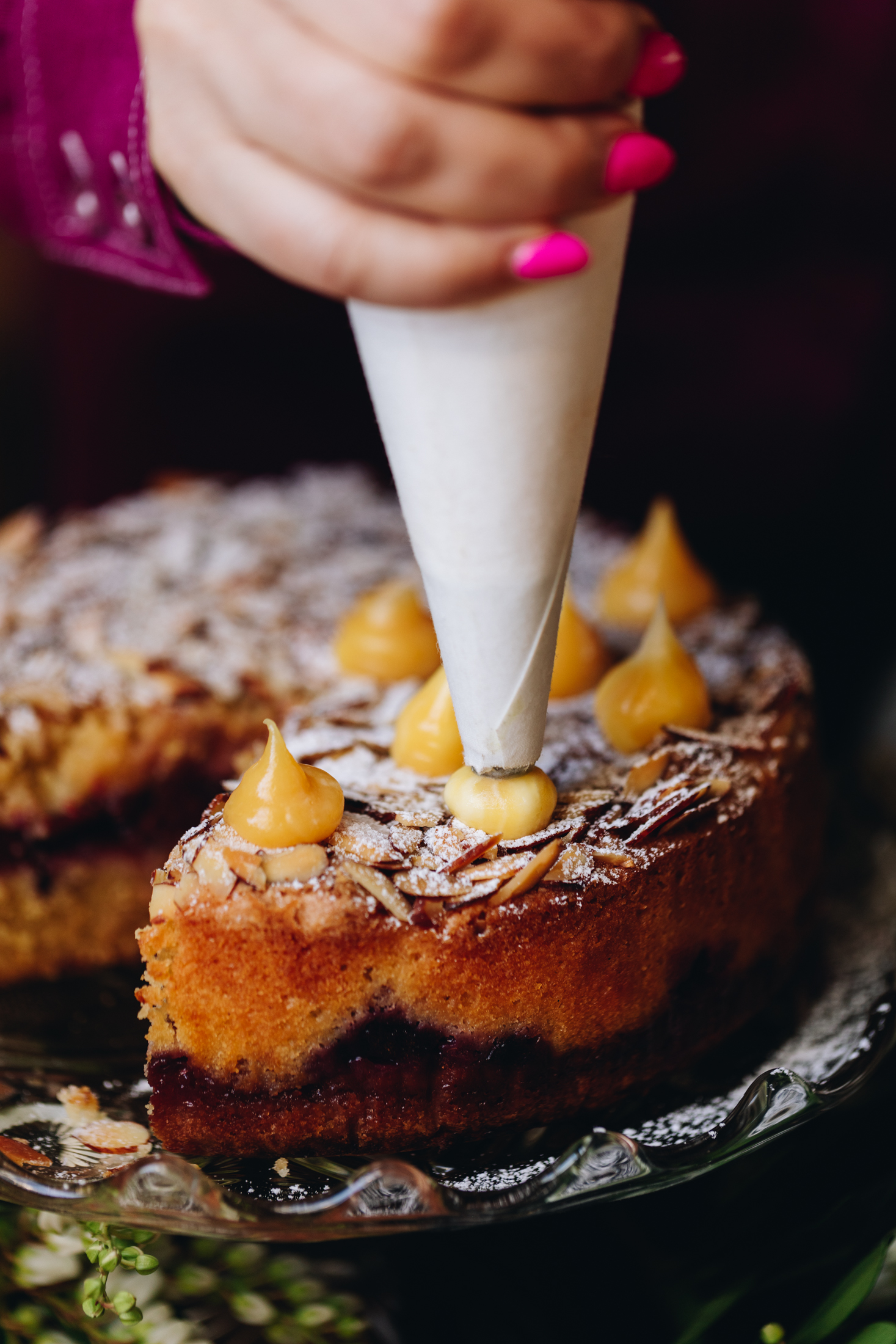On a glass cake stand sits a freshly baked plum and almond cake. It has been dusted with icing sugar and is being piped with creamy lemon curd.