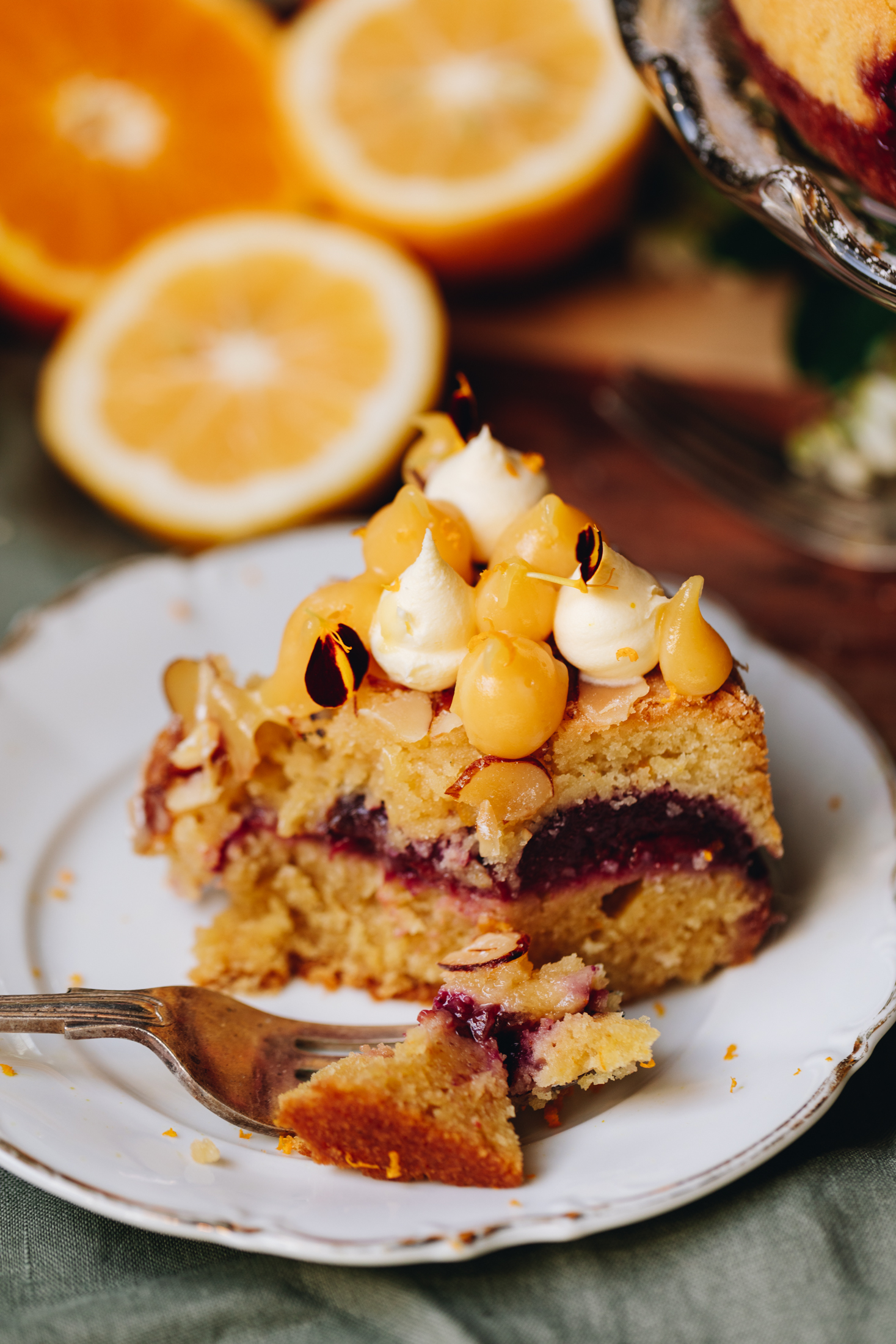 On a white plate with gold trim sits a slice of plum and almond cake that has been dug in to with a fork. It shows the three layers of orange cake, black Doris plums and almond cream with a topping of lemon curd and mascarpone piped on top. Cut oranges are in the background.