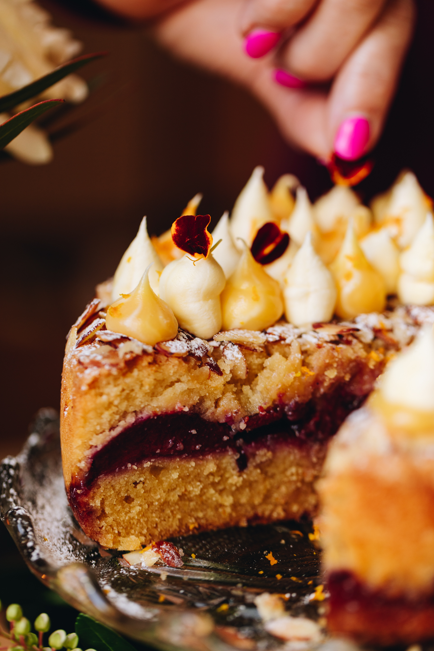 On a glass cake stand sits a freshly baked plum and almond cake. It has been dusted with icing sugar with a topping of lemon curd and mascarpone piped on top. It is being topped with a flower petals. The cake has been cut open to reveal the three layers of orange cake, black Doris plums and almond cream.