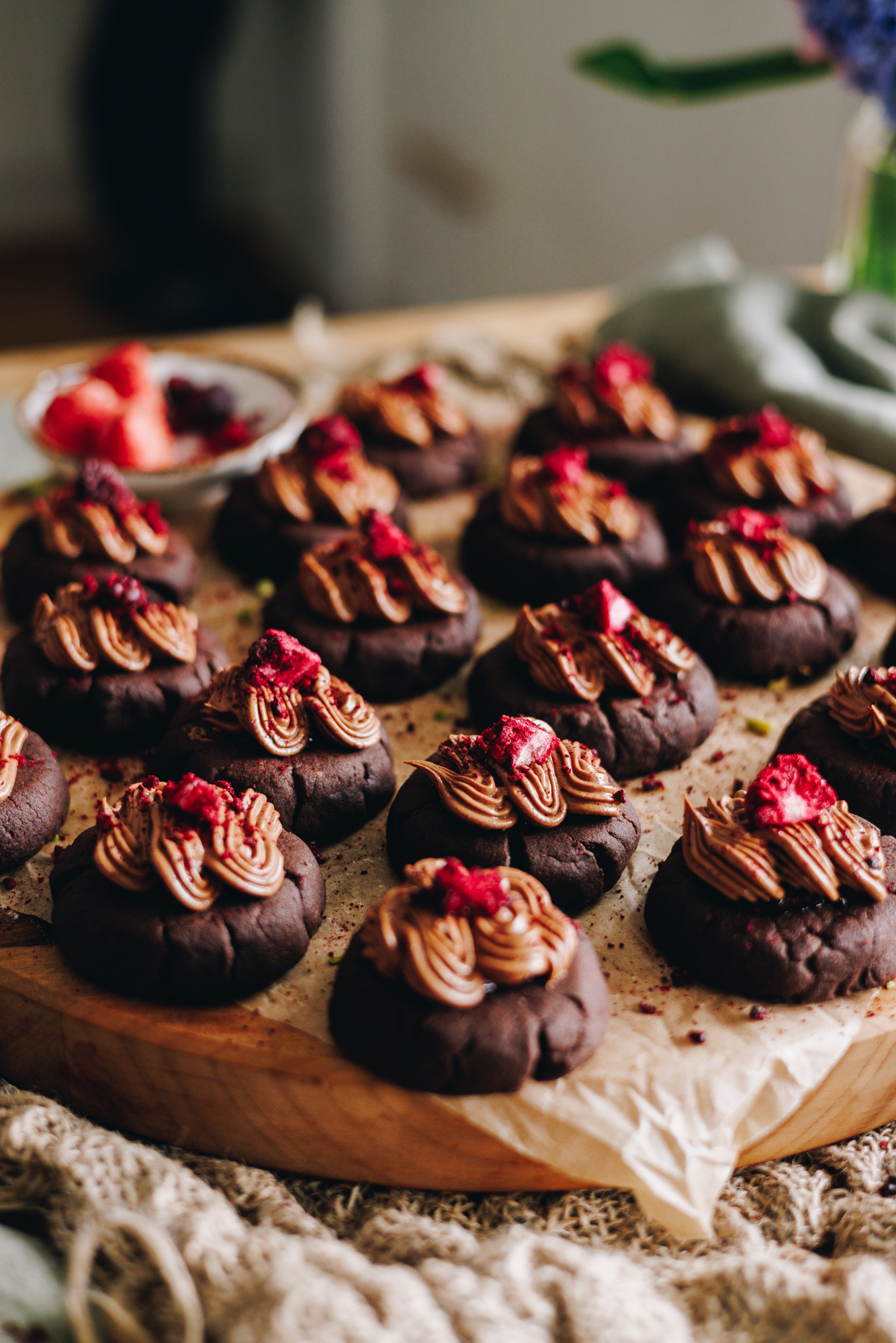 On a table covered in natural fabrics is a wooden board that is covered in brown baking paper. On top are decorated chocolate and raspberry cookies. They have chocolate frosting on top of each one and freeze-dried boysenberries sit on top. 