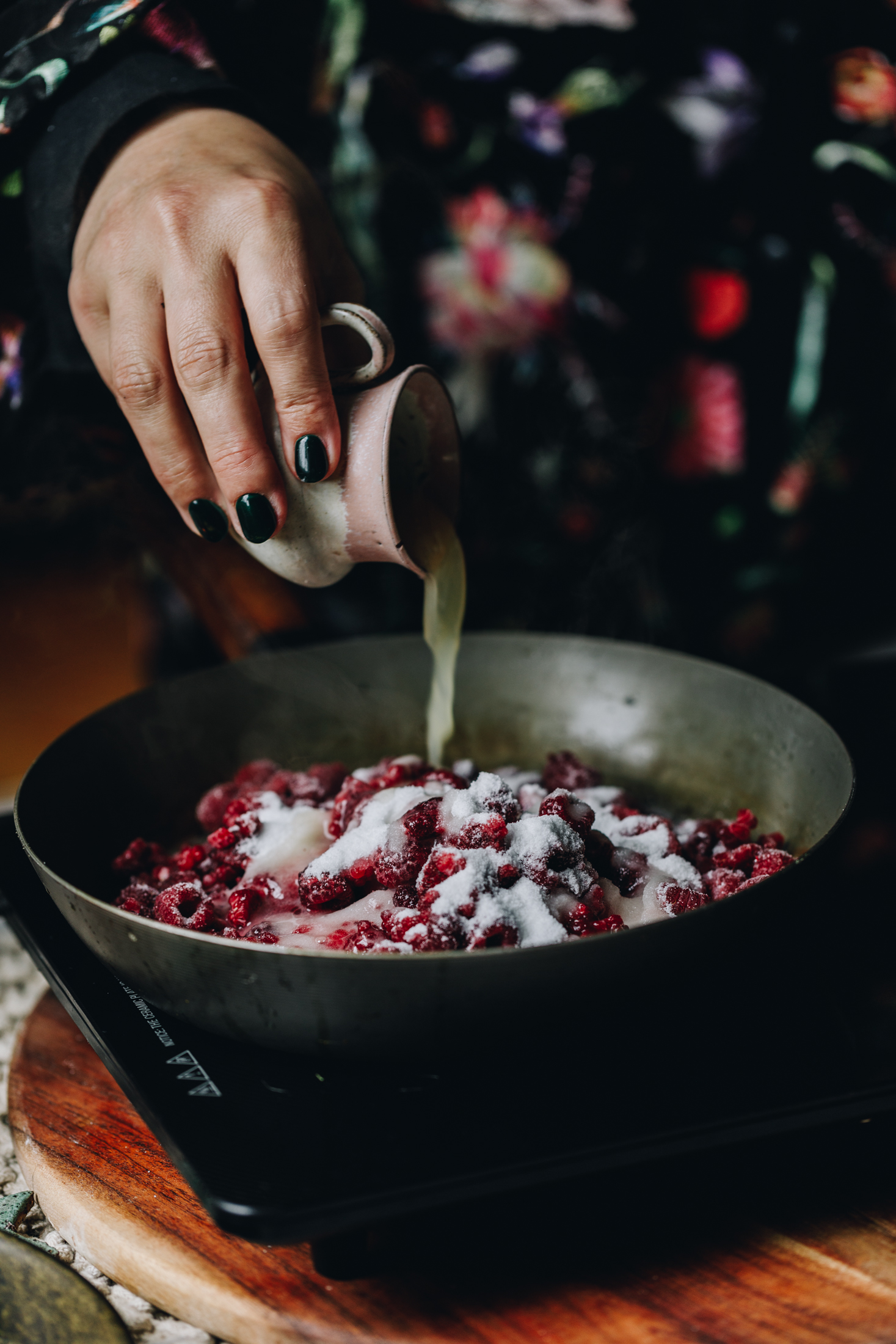On a wooden table is a black portable stove and a cast iron pan sits on top of it. in the pan is frozen raspberries and sugar. A small pink ceramic jug is pouring lemon juice in to it. 