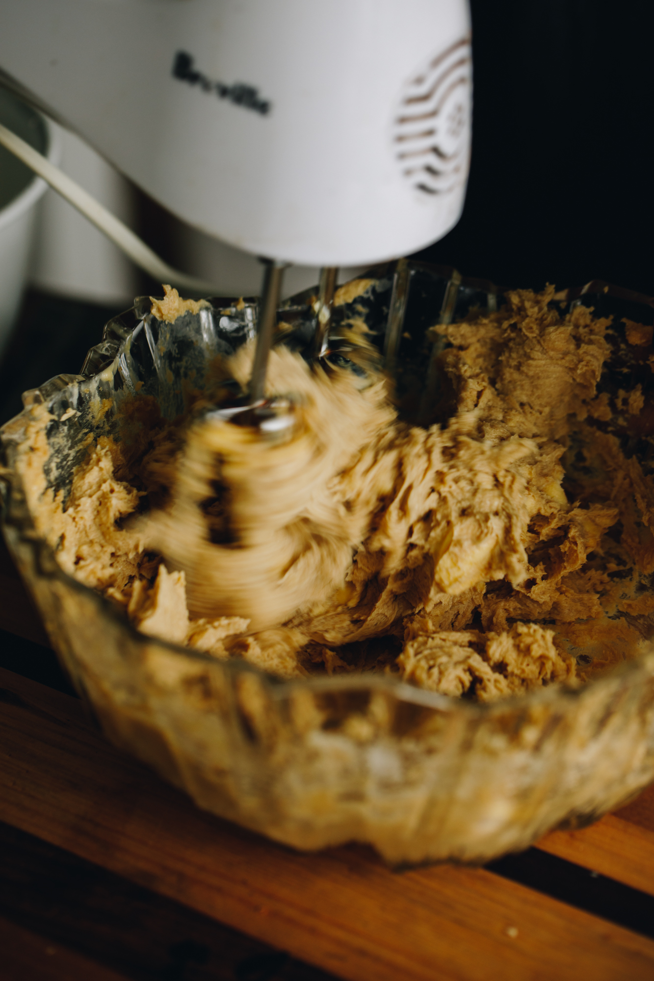 In a vintage glass bowl is butter and sugar being whipped with a w white hand mixer. It is sitting on a wooden board.