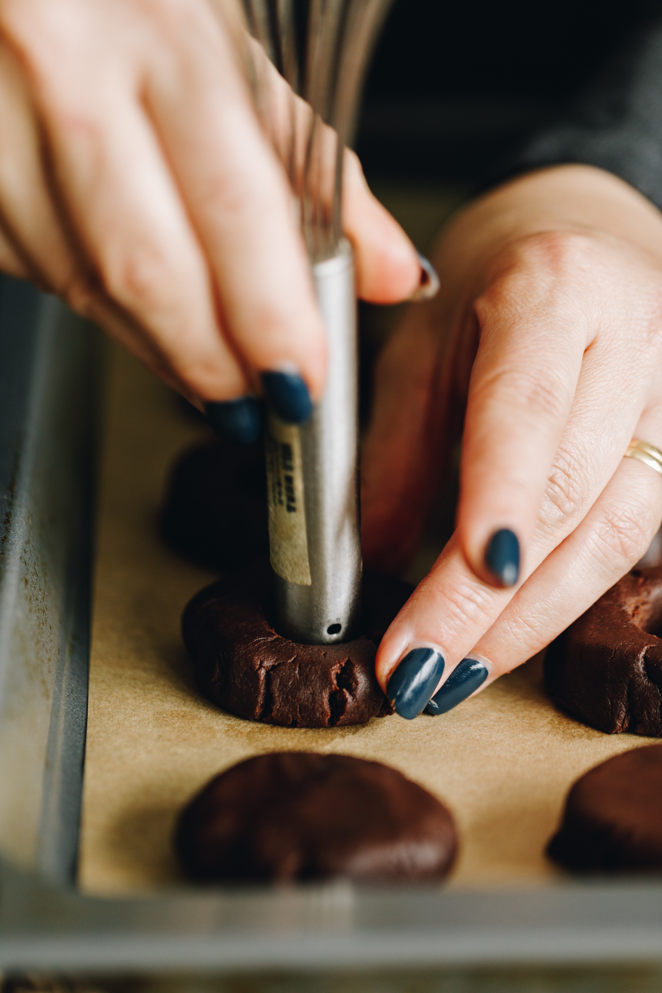 On a tray with brown baking paper are unbaked chocolate cookies that have been flattened down. Naomi is indenting one with a silver handle.
