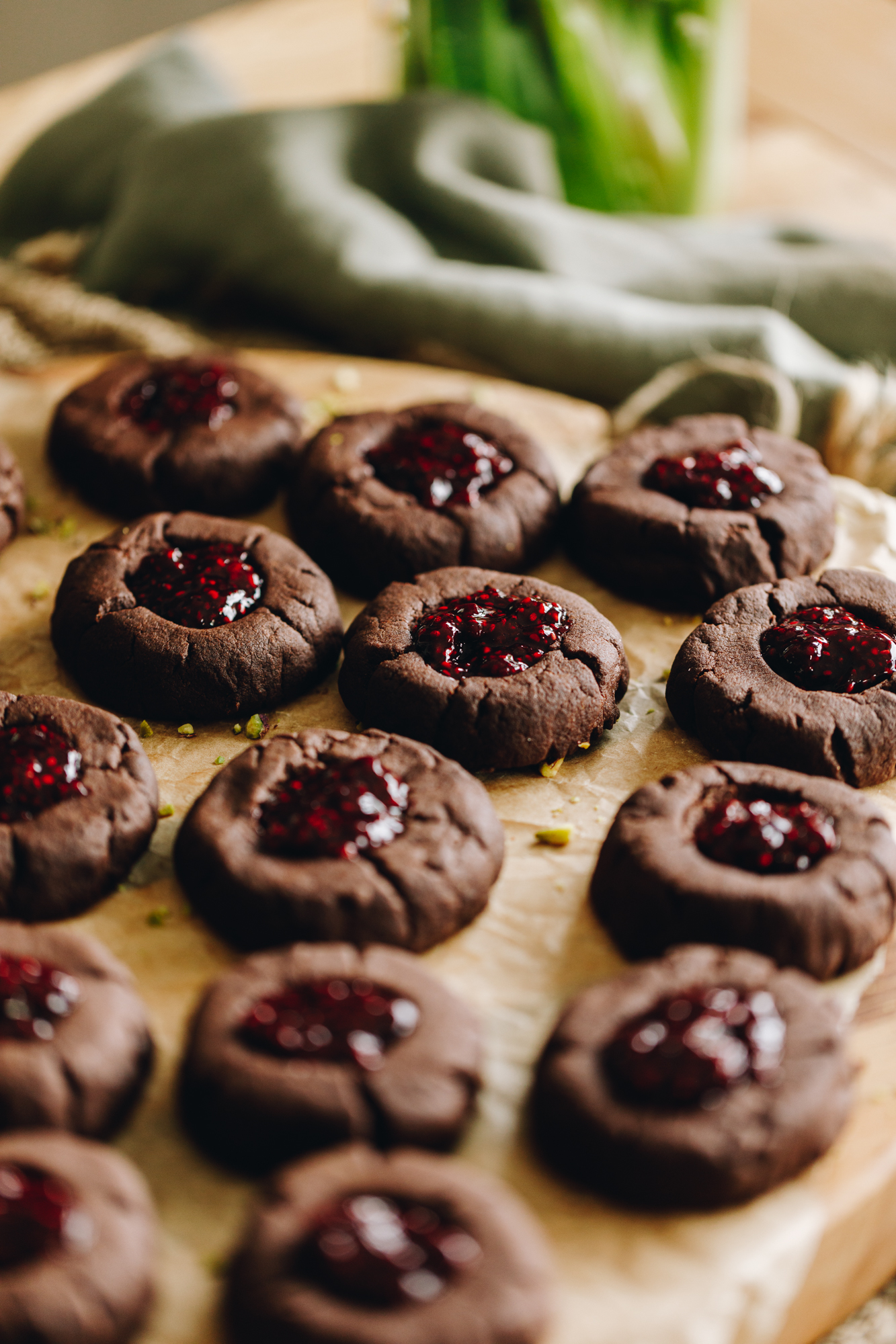 On a board with brown baking paper sits freshly baked chocolate and raspberry cookies that have been filled with raspberry jam. A green tablecloth is ruffled in the background.