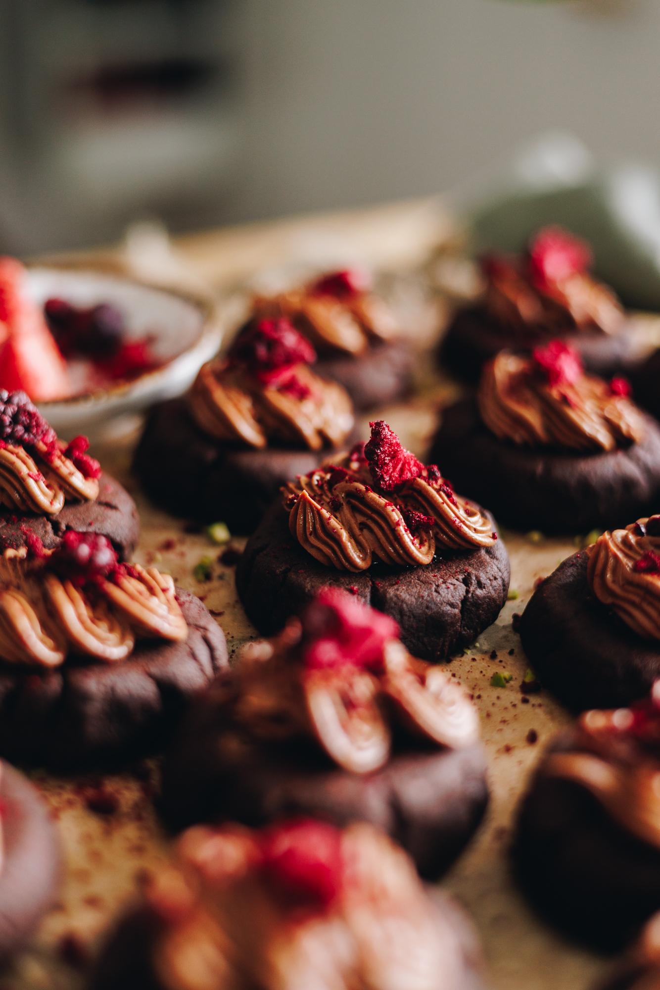 A wooden board is covered in brown baking paper. On top are decorated chocolate and raspberry cookies. They have chocolate frosting on top of each one and freeze-dried boysenberries sit on top. 