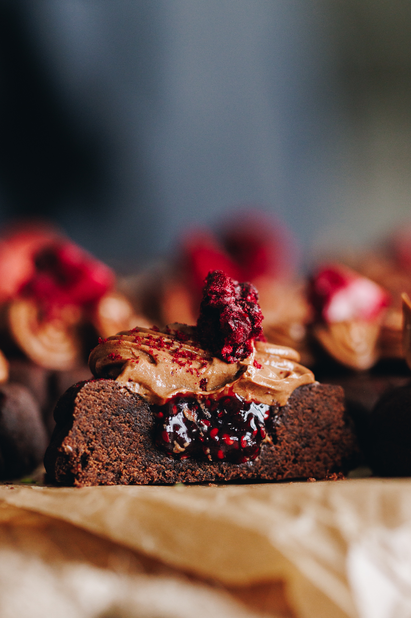 A close up shot shows a board covered in brown baking paper. On top are decorated chocolate and raspberry cookies. One has been cut open revealing the jam inside and soft texture of the buttercream. 