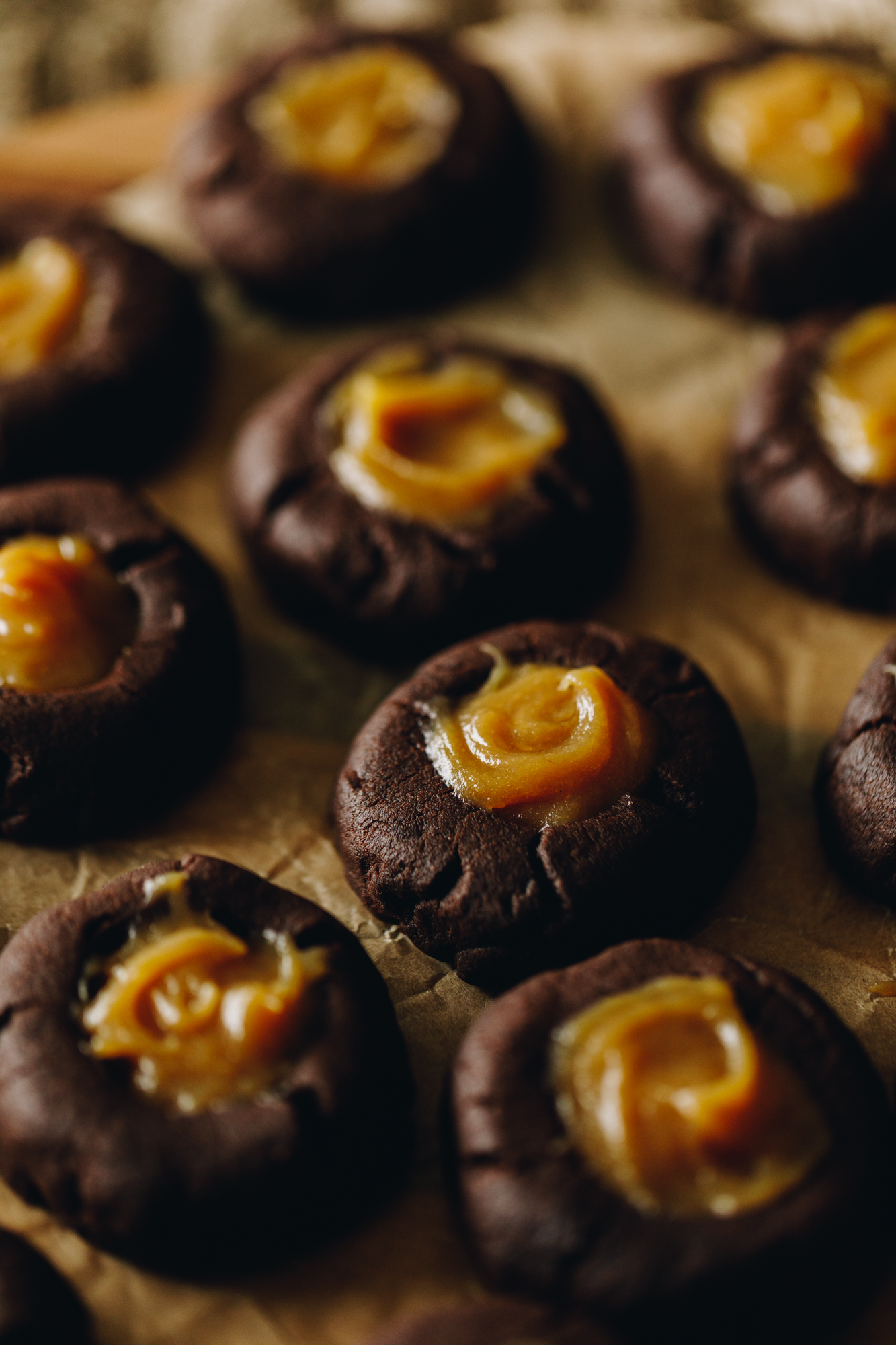 On a board with brown baking paper sits freshly baked caramel filled chocolate cookies that have been filled with thick caramel. 