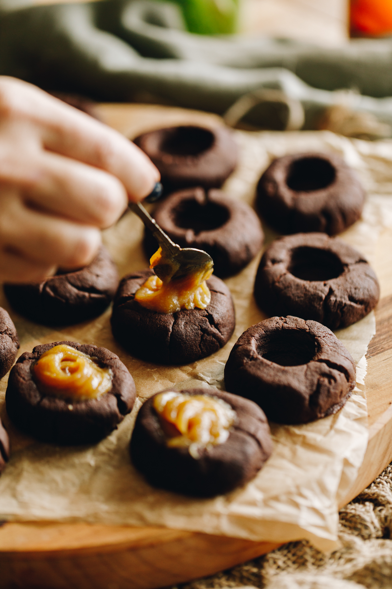 On a board with brown baking paper sits freshly baked caramel filled chocolate cookies that are being filled with caramel. Some cookies have nothing in them and three have caramel. A green tablecloth is ruffled in the background.