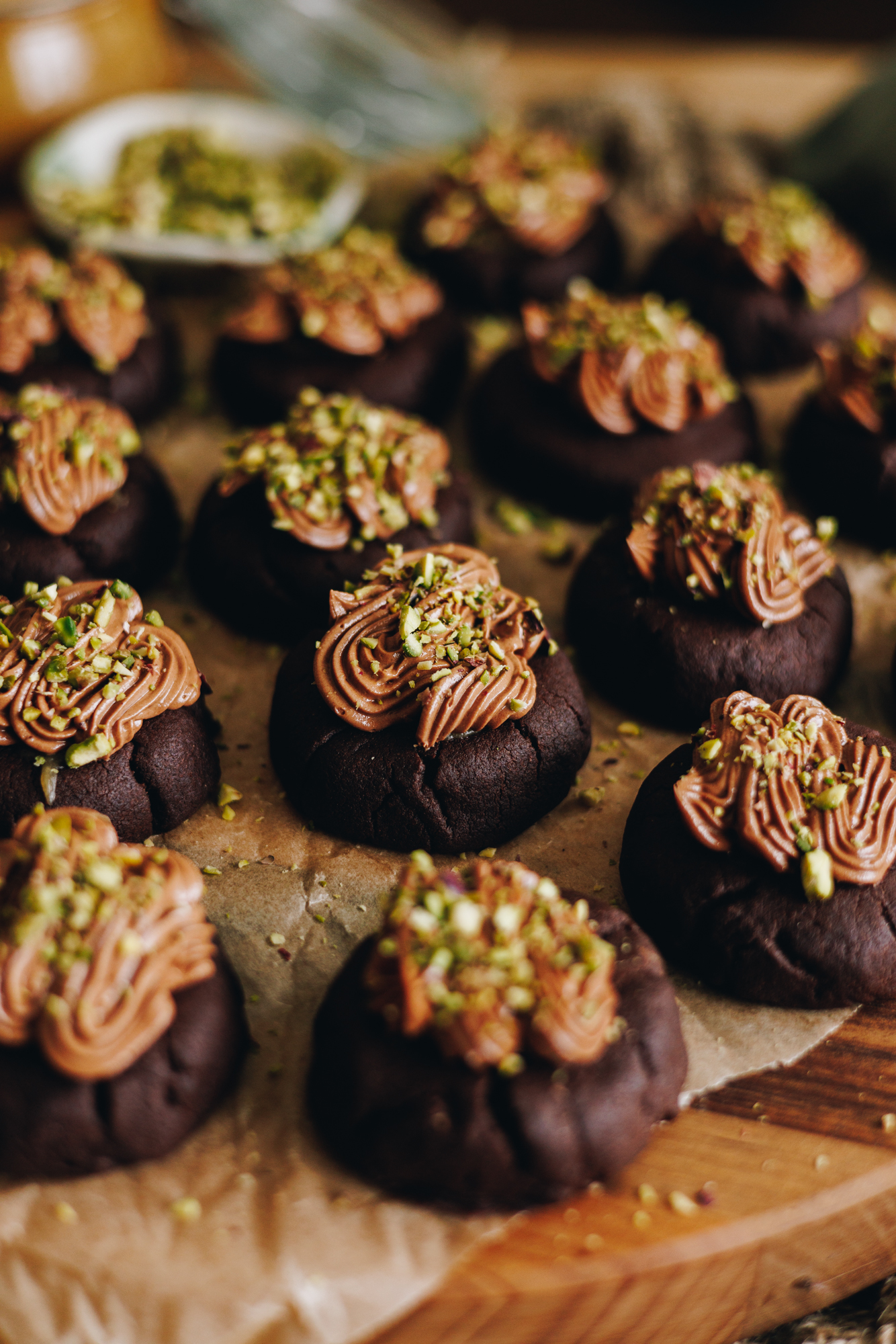 A wooden board that is covered in brown baking paper sits on a table. On top are decorated caramel filled chocolate cookies. They have chocolate frosting on top of each one and crushed pistachios sit on top.