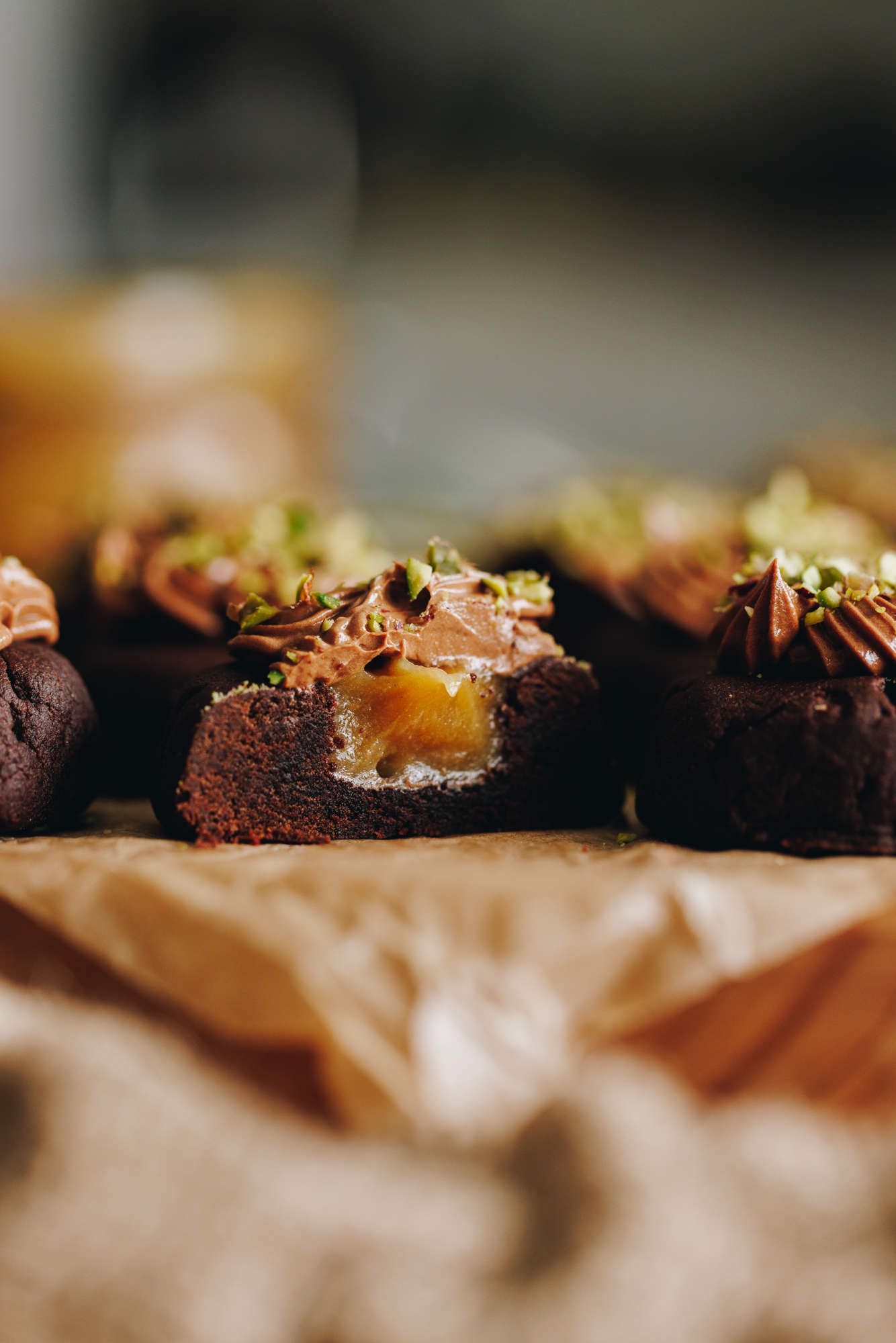 A close up shot shows a board covered in brown baking paper. On top are decorated caramel filled chocolate cookies. They have chocolate frosting on top of each one and chopped pistachios sit on top. One biscuit has been cut in half to expose the caramel inside. 